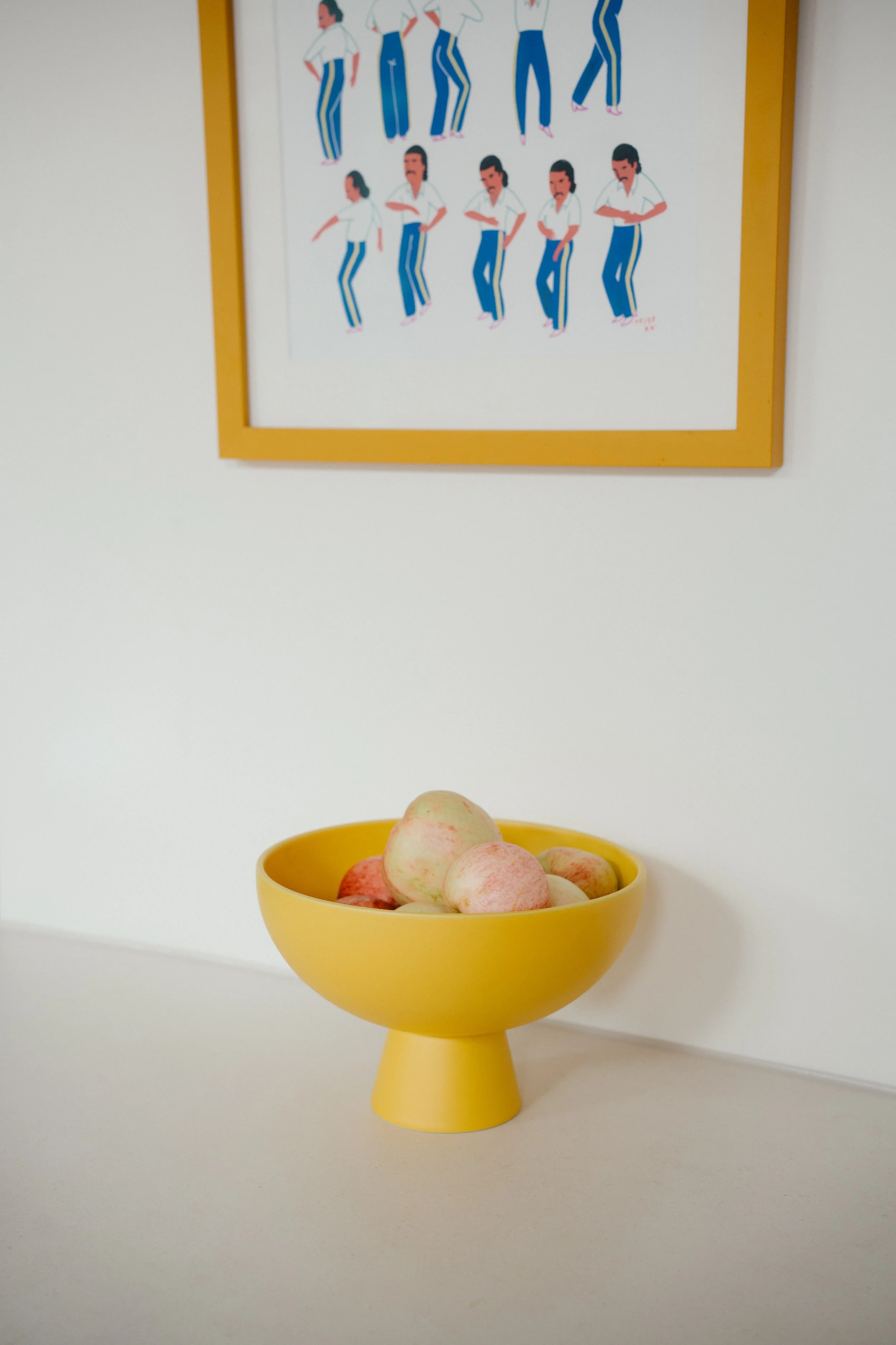A yellow bowl filled with light-colored apples sits on a white surface. Behind it, on the wall, is a framed illustration of multiple figures dressed in white shirts and blue pants, depicted in various poses.