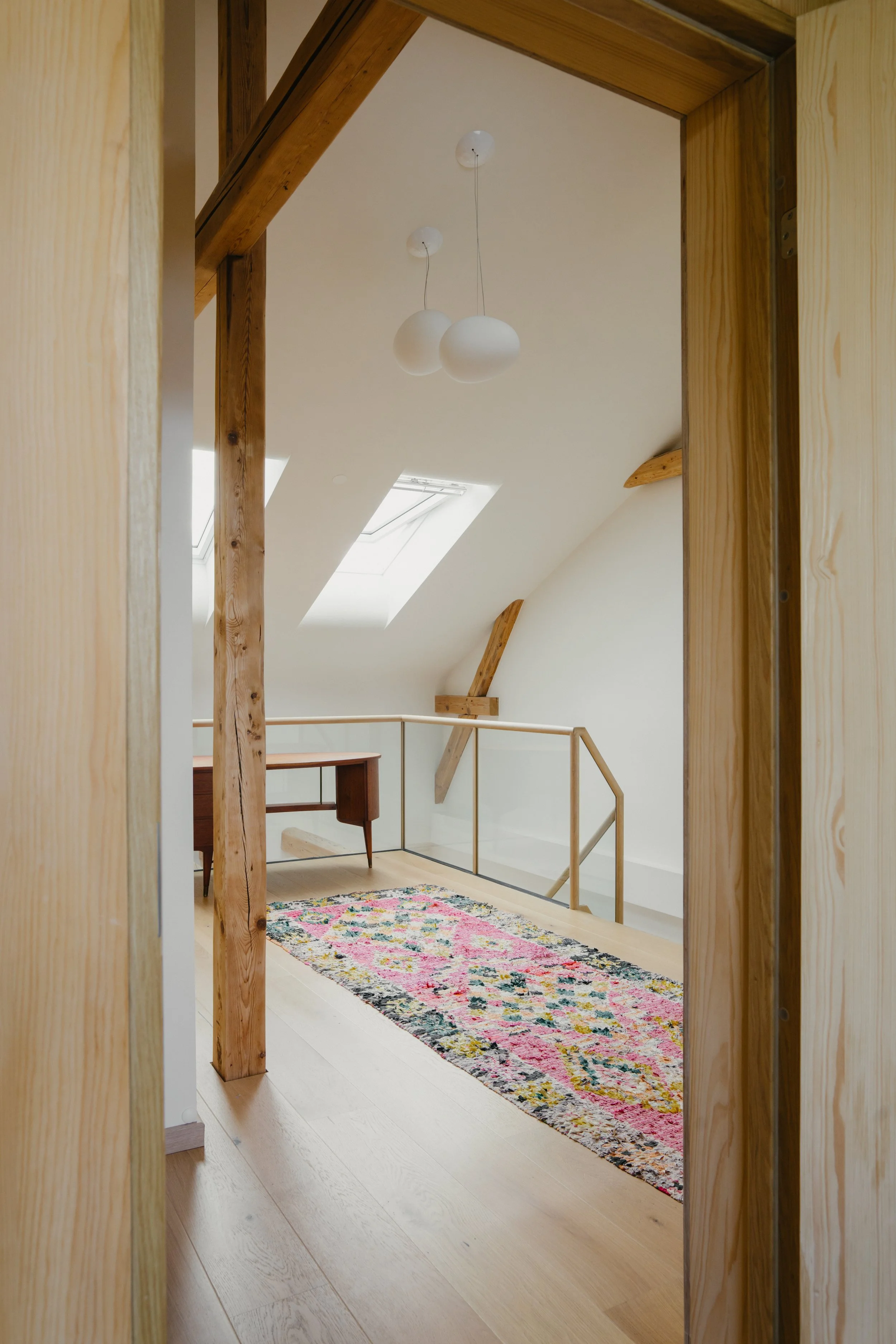 Interior view of a room with wood beams, a skylight, a hanging pendant lamp, a colorful rug, a small wooden desk, and a staircase railing.