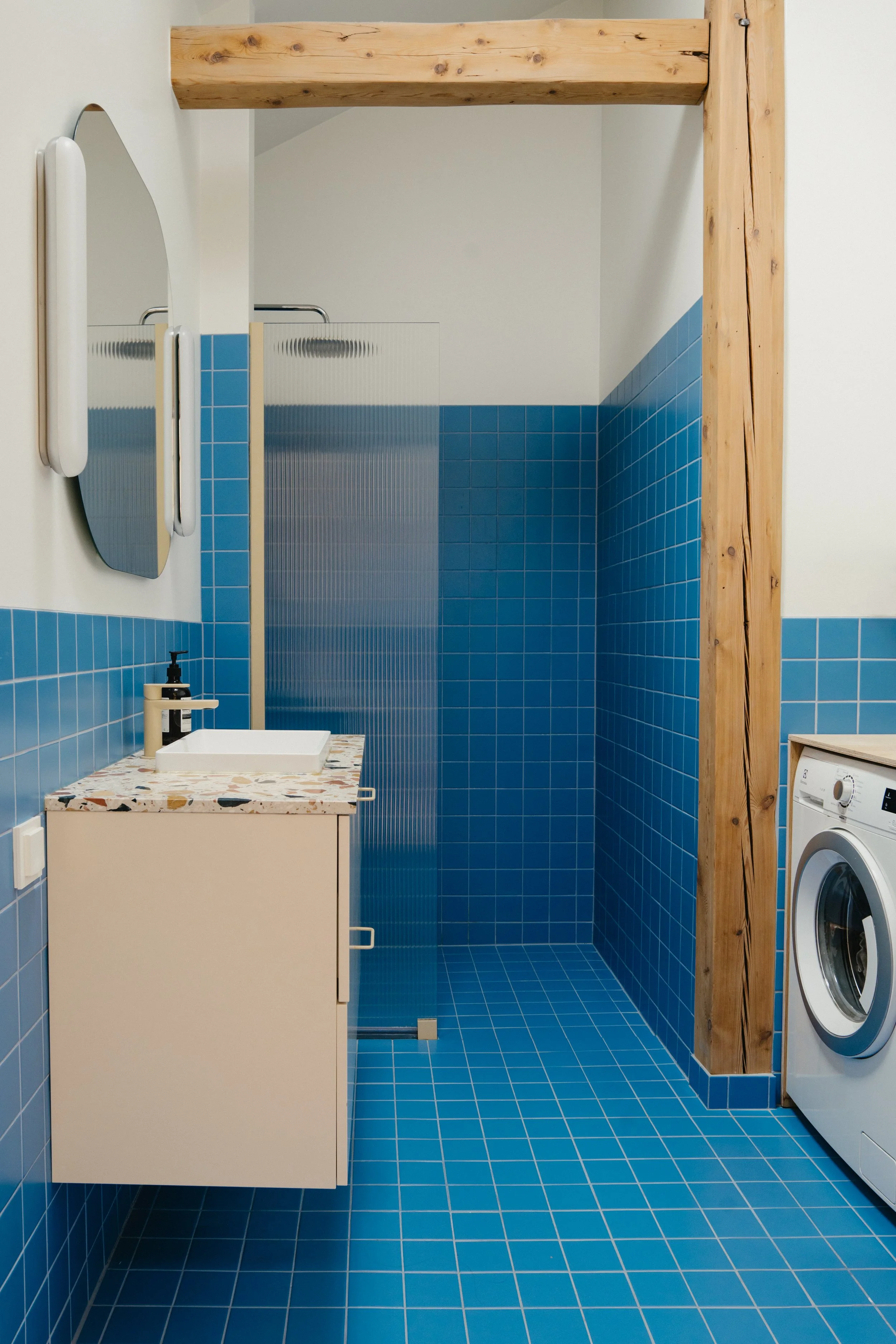 Modern bathroom with blue tile walls and floor, a white sink on a beige cabinet with a speckled countertop, a mirror, a washing machine, and a shower with a glass and textured panel, framed by a wooden beam.