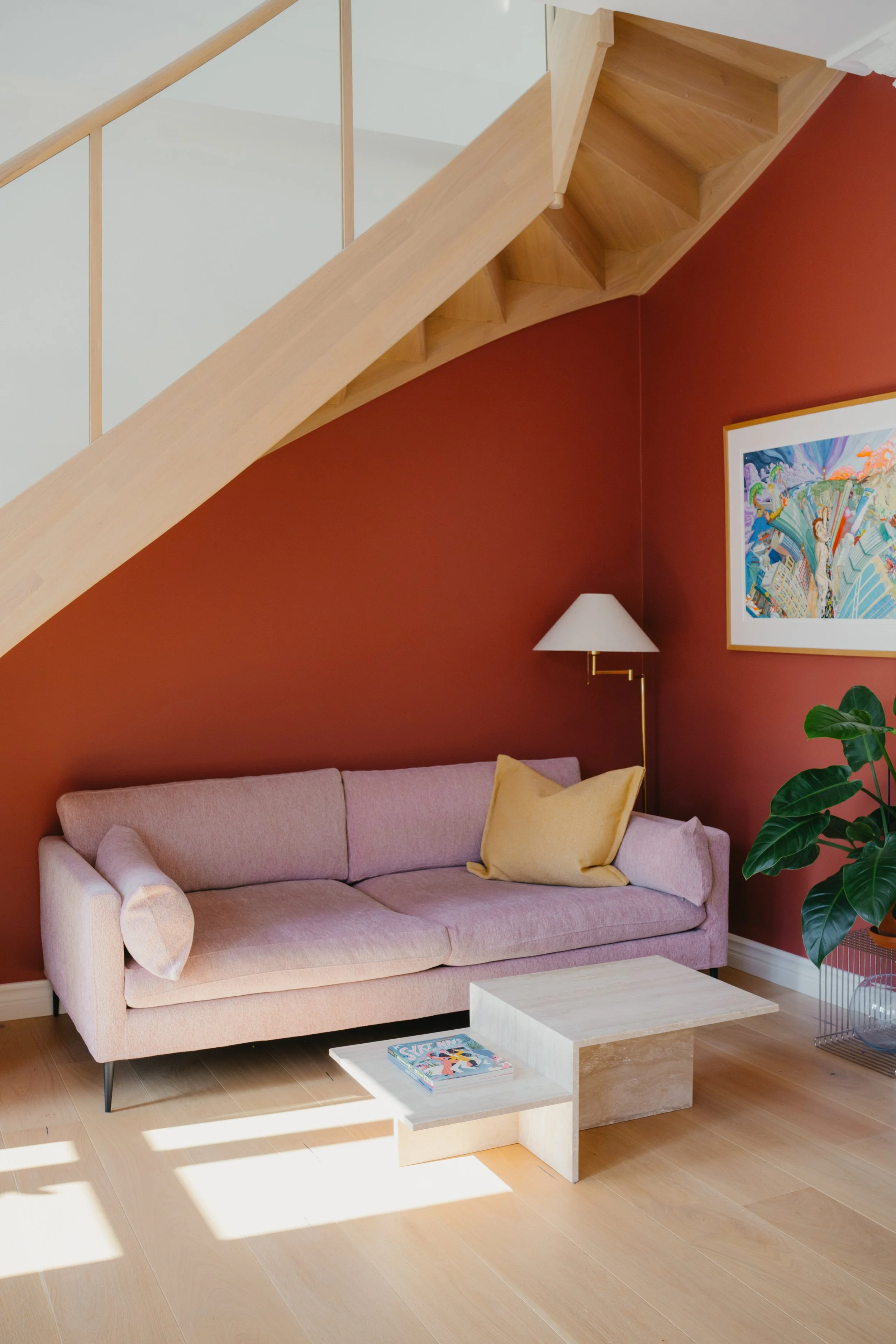 Interior of a living room with a pink sofa, a beige coffee table, a yellow pillow, a white wall-mounted lamp, colorful artwork on a red wall, a large green plant, and a wooden staircase overhead.