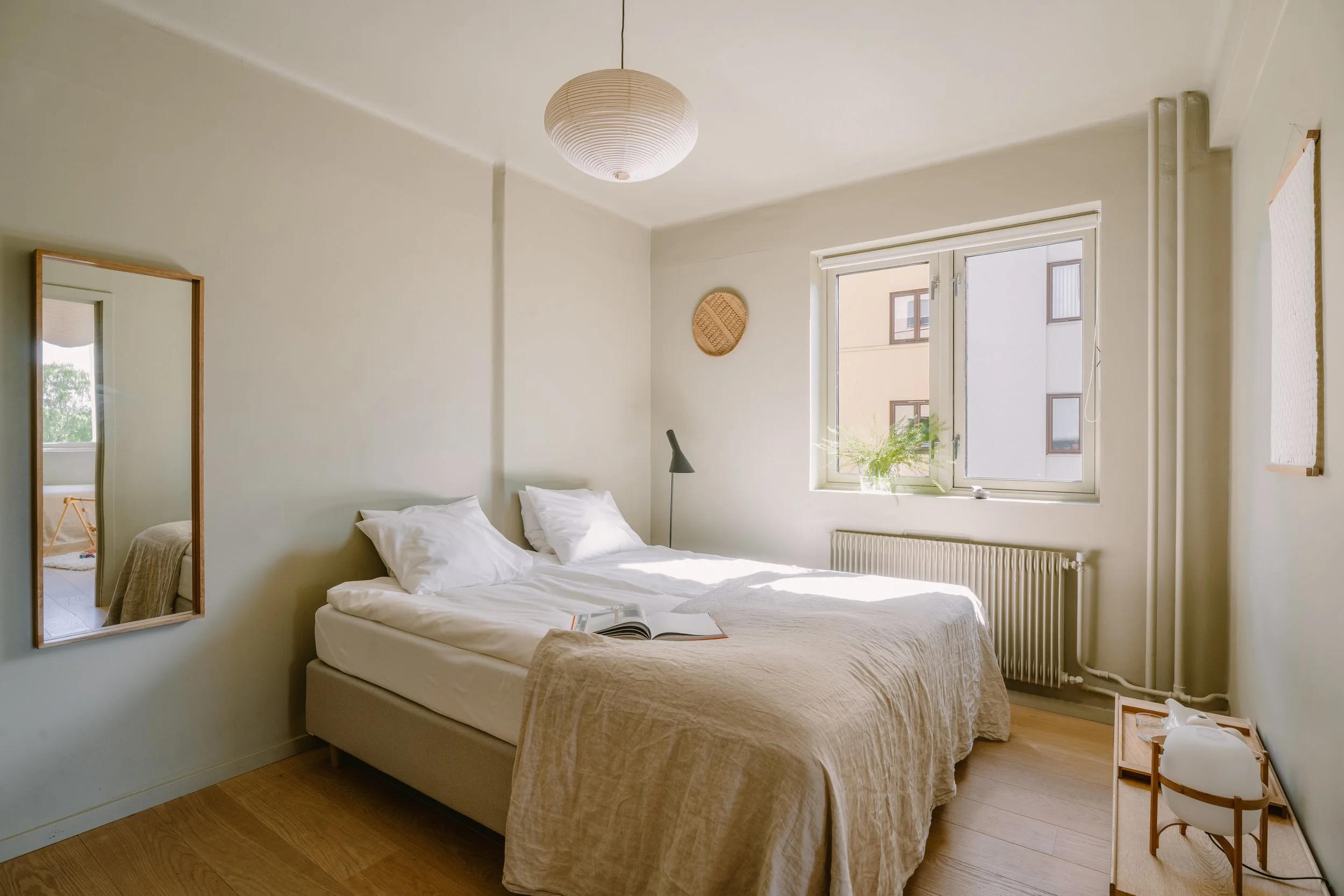 A bright bedroom with a large bed, white bedding, and pillows. A mirror on the wall reflects part of the room. A window lets in natural light, with a small plant on the windowsill. A ceiling light fixture and a wall-mounted light are visible.