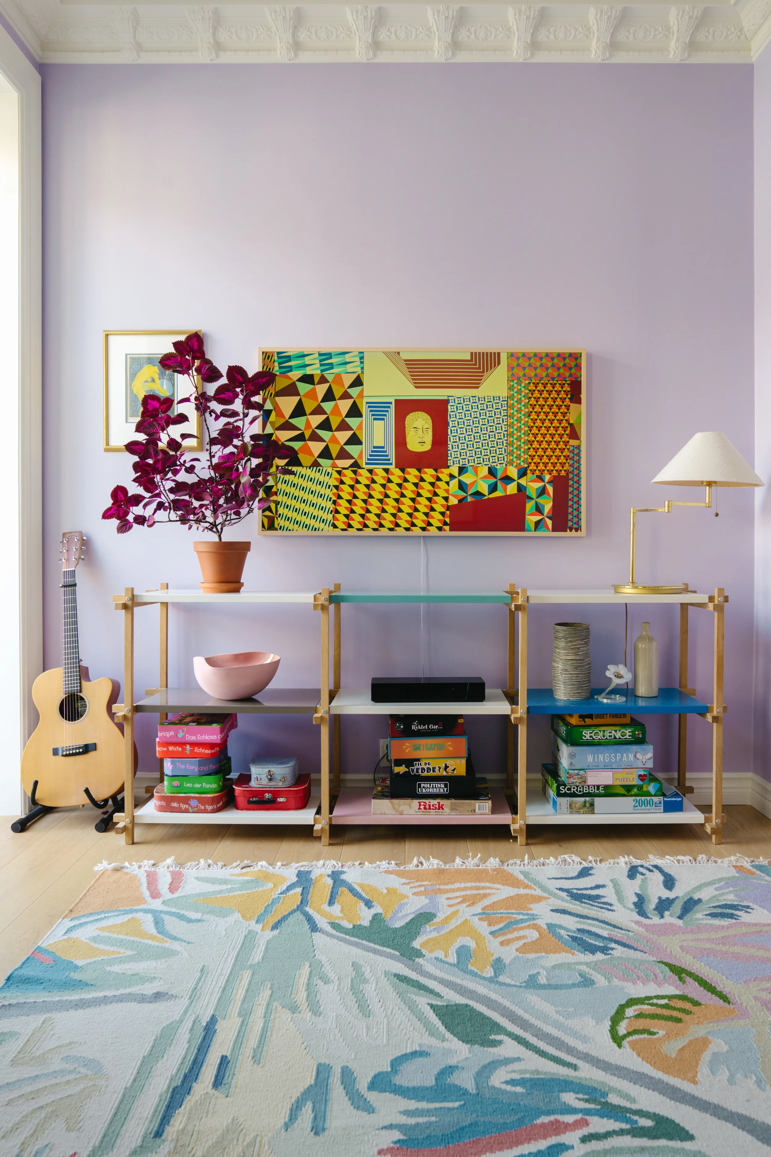 A colorful living room corner with a purple wall, a wooden shelving unit holding books, decorative objects, and a lamp. A guitar leans against the wall on the left, and a rug with multicolored patterns is on the floor. Artwork hangs on the wall above