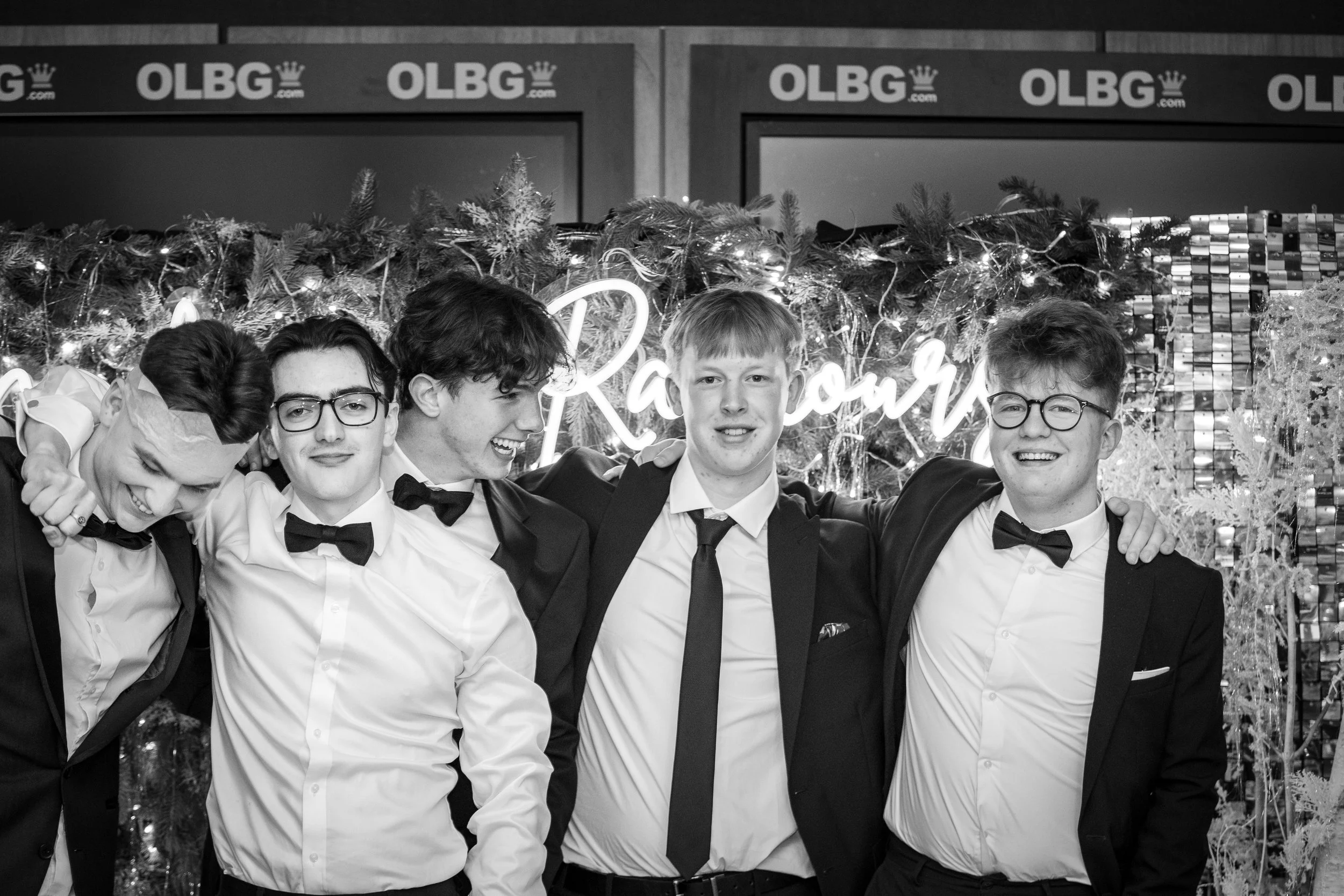 A group of five young men dressed in formal tuxedos and bow ties, standing close together with their arms around each other's shoulders, smiling and looking happy. They are at a festive event with holiday decorations, including garland with lights an