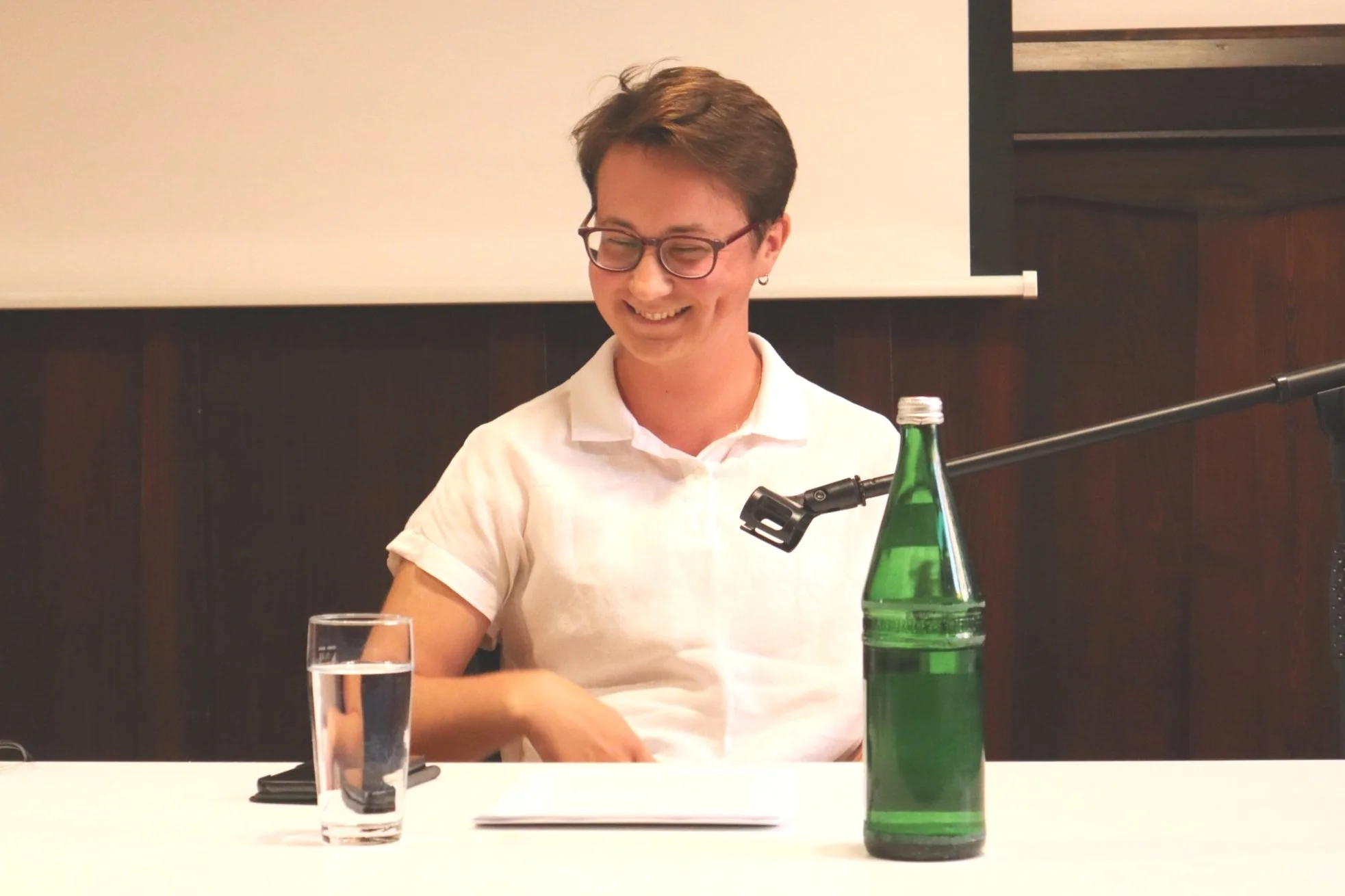 Eine lächelnde Person sitzt an einem Tisch mit einem Mikrophon, einem Glas Wasser, einer grünen Glasflasche und einem Papierstapel.
