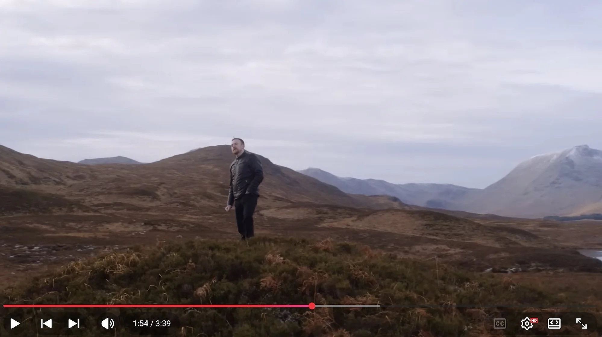 A man stands alone in a vast, open landscape with rolling hills and distant mountains under a cloudy sky.