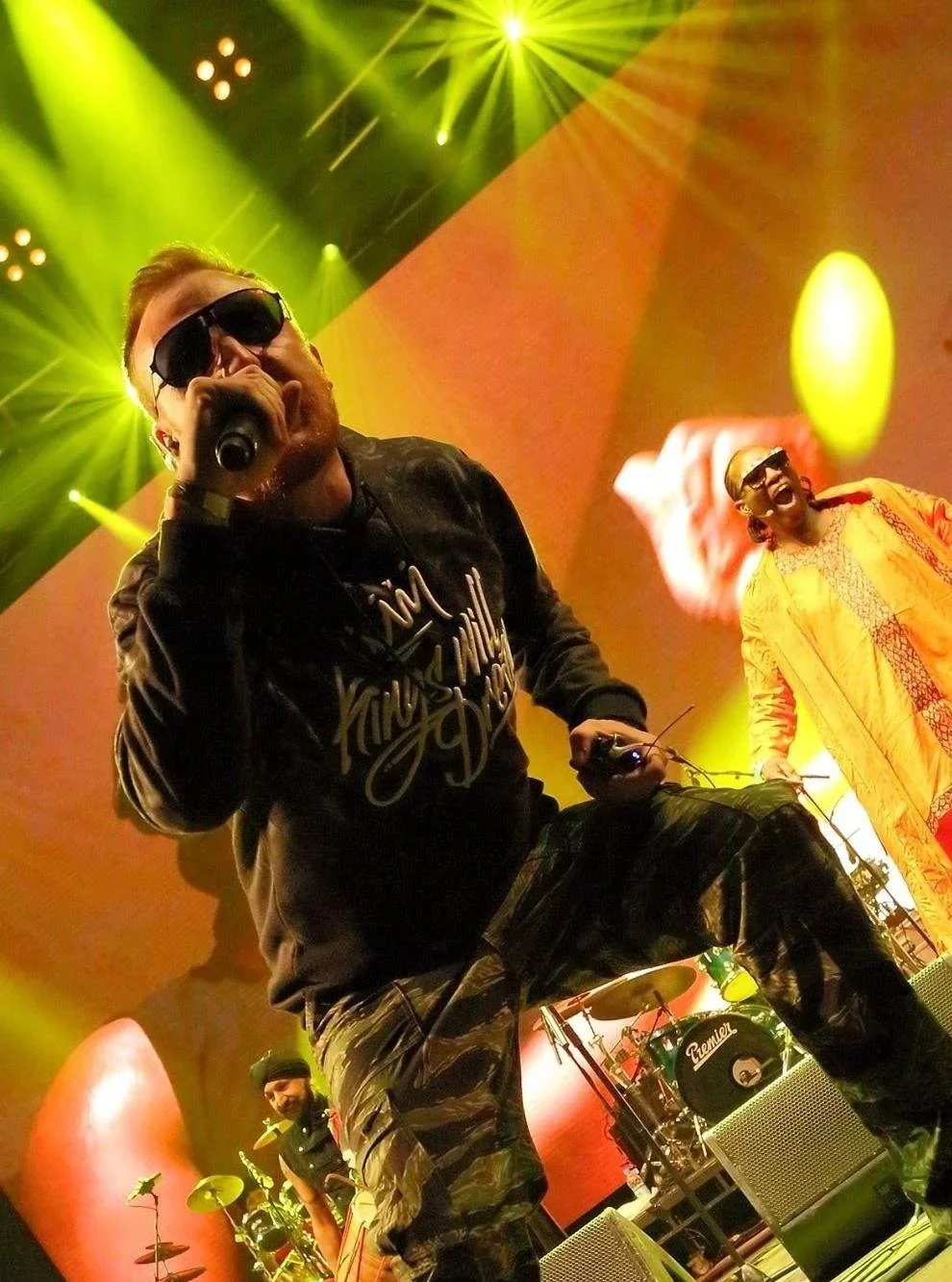 Griogair Labhruidh performing live with Afro Celt Sound System in 2016. He’s rapping, front and centre in sunglasses and a black sweatshirt, crouching toward the camera with a mic in hand.