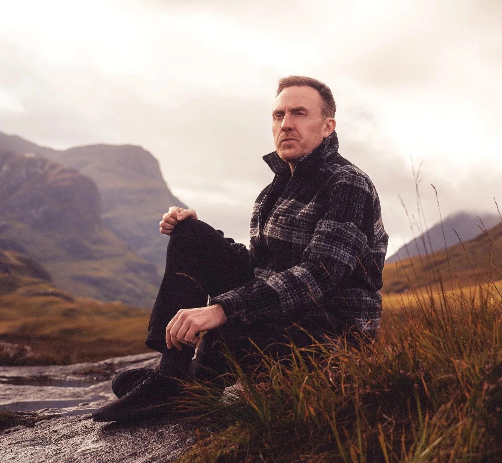 Scottish Gaelic singer Griogair Labhruidh seated in the Glencoe landscape beneath dramatic Highland skies.