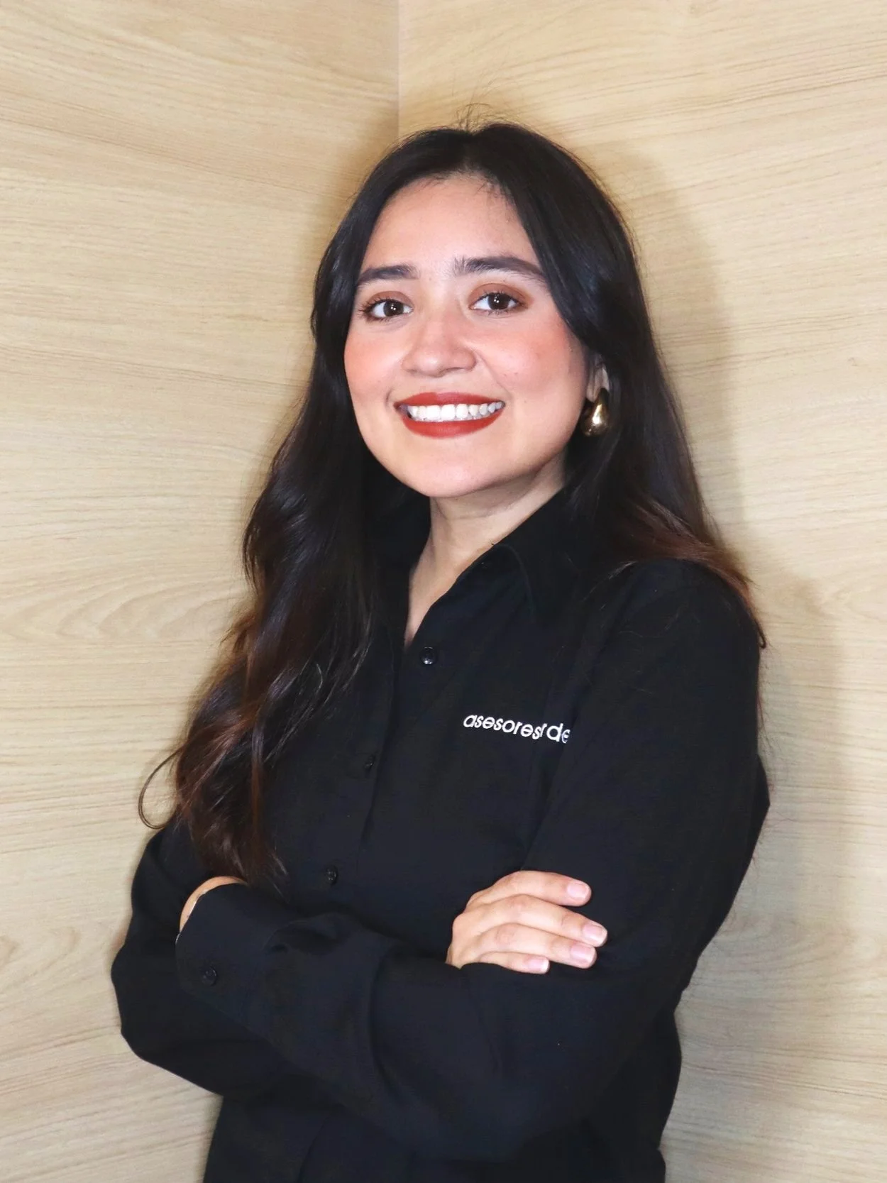 Una mujer de cabello oscuro y largo, sonriendo, vestida con una camisa negra que tiene texto en el lado izquierdo del pecho, posando de pie con los brazos cruzados frente a una pared de madera clara.