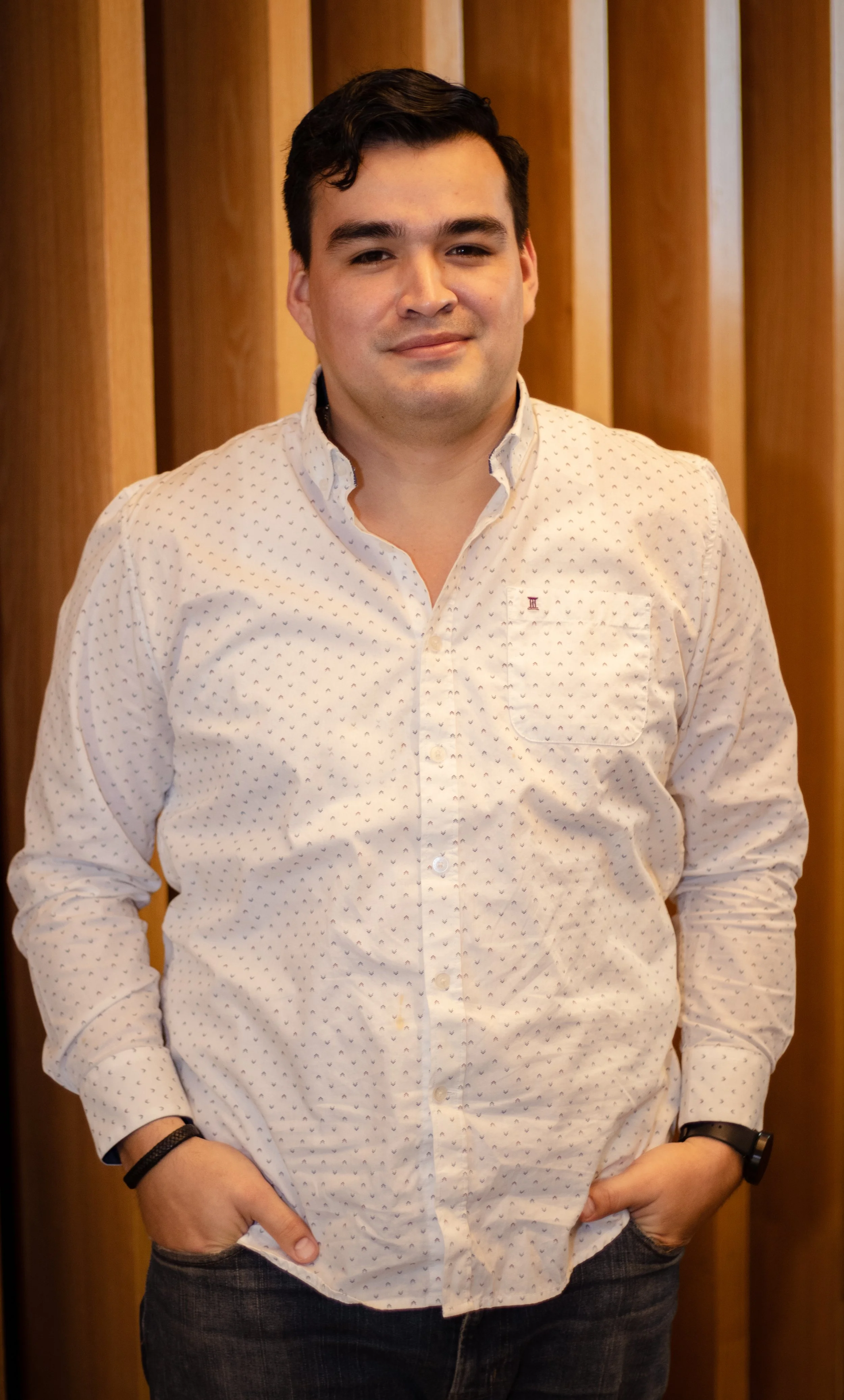 Persona con camisa blanca de puntos, manos en los bolsillos, de pie frente a un fondo de listones de madera verticales.