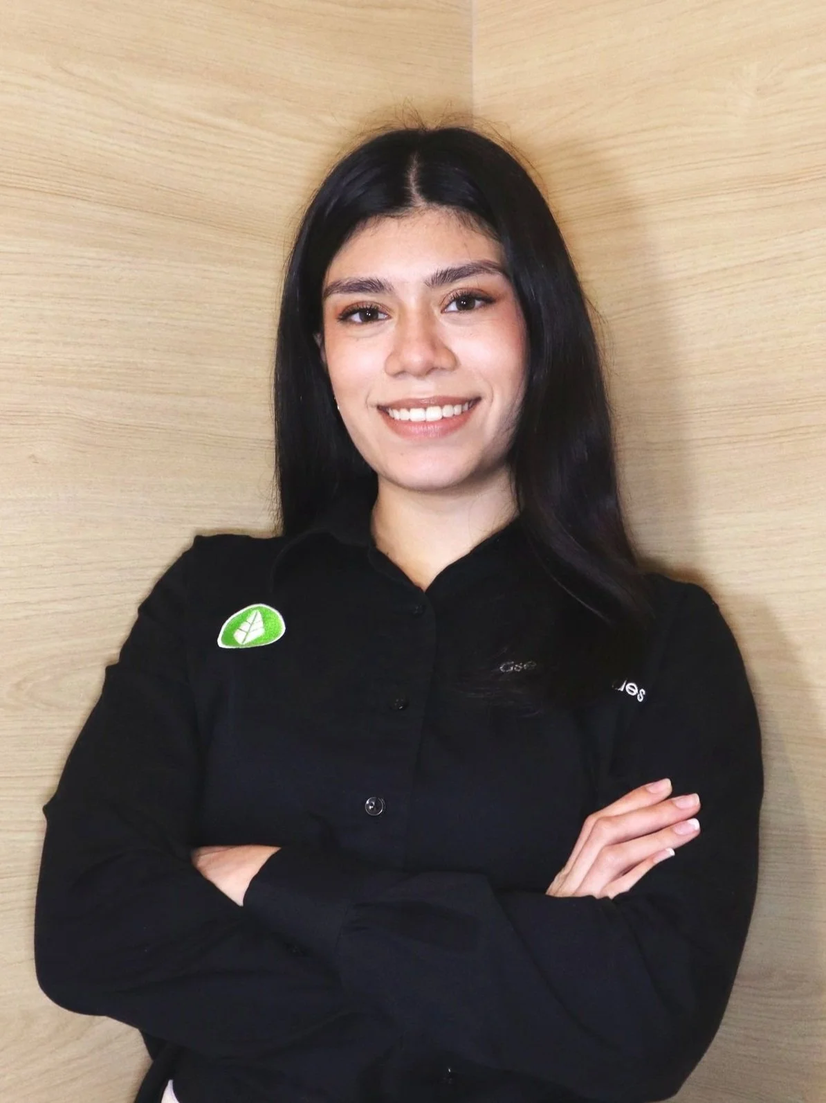 Mujer sonriendo con camisa negra, brazos cruzados frente a pared de madera clara.