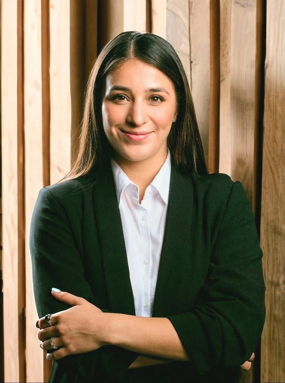 Mujer joven con cabello largo, vistiendo un traje formal negro y camisa blanca, sonríe con los brazos cruzados frente a un fondo de madera.
