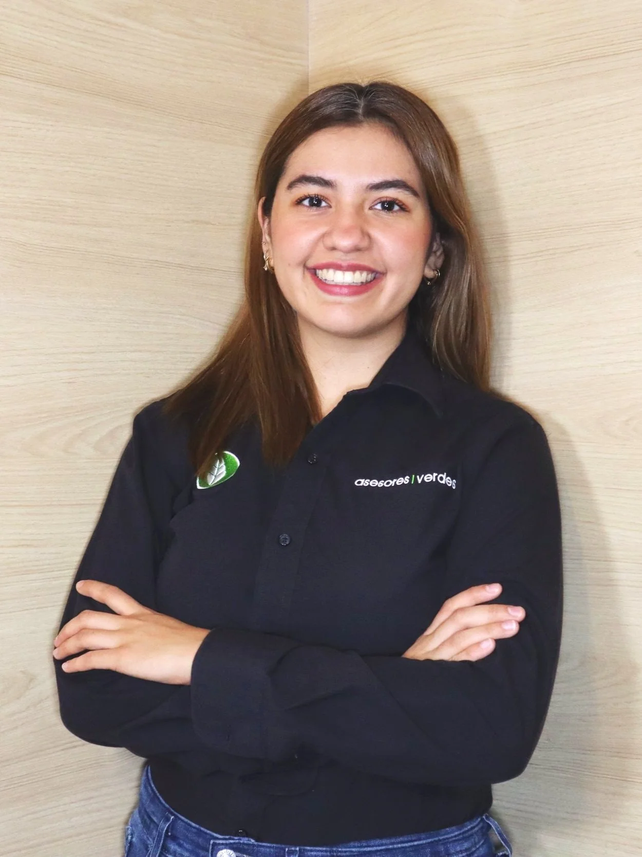 Mujer joven sonriendo con camisa negra, brazos cruzados, fondo de madera clara.