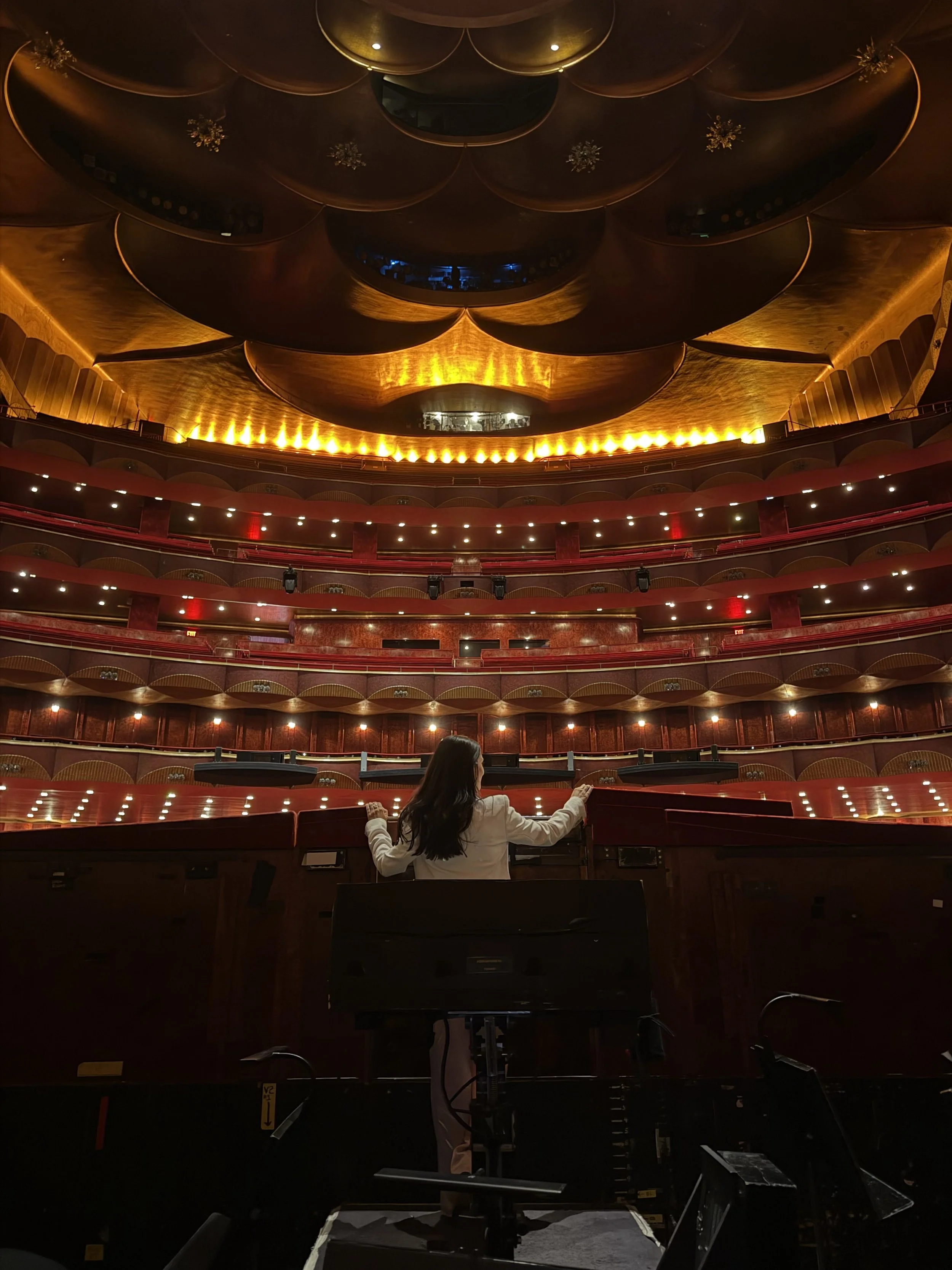 The image shows the interior of a large theater with an ornate ceiling lit by golden lighting. There are multiple balconies with red railings, and a woman dressed in white is sitting at the front, facing the stage. The stage is not visible in the image.