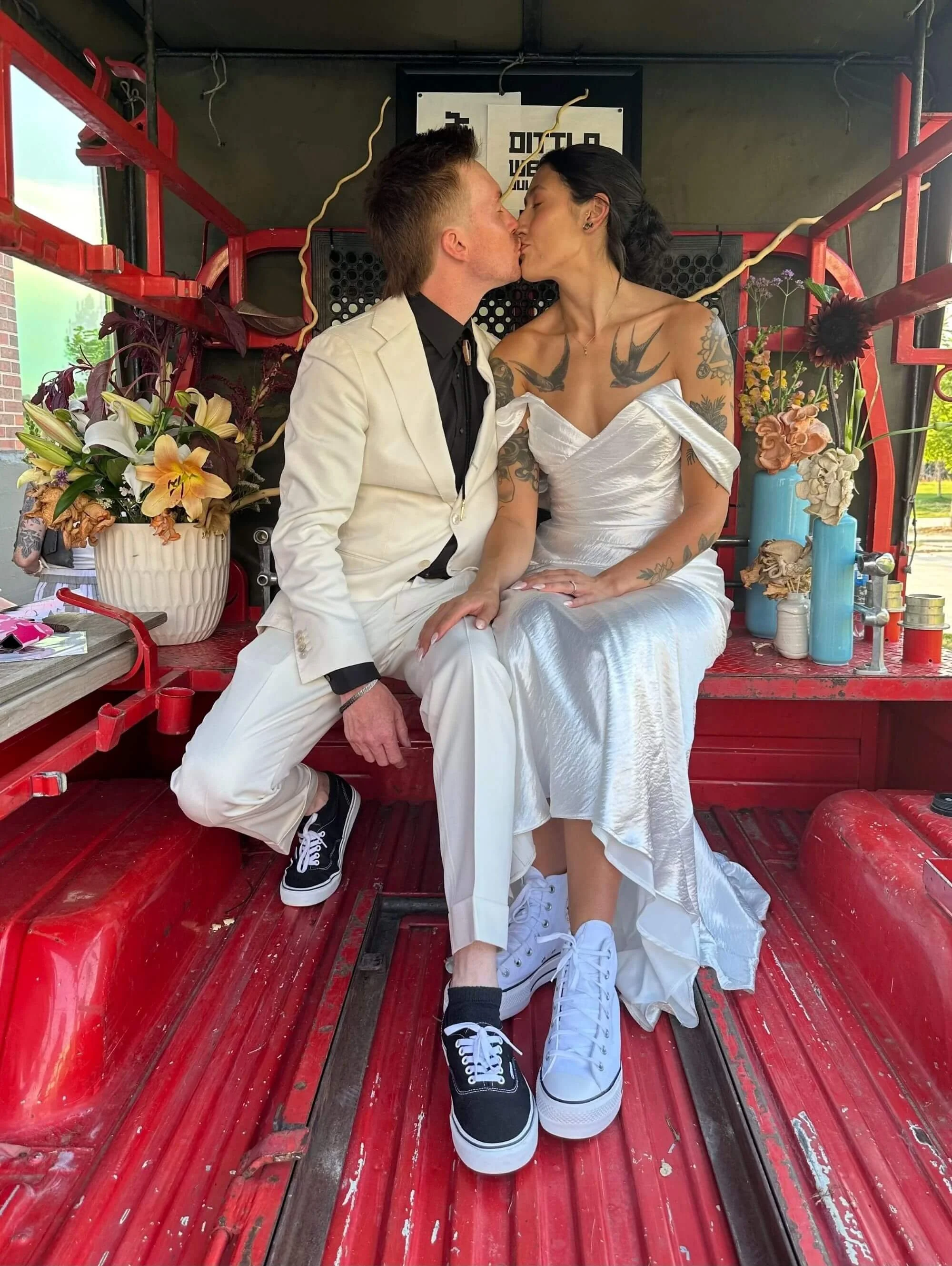 A couple in wedding attire kissing while seated among seasonal floral arrangements.