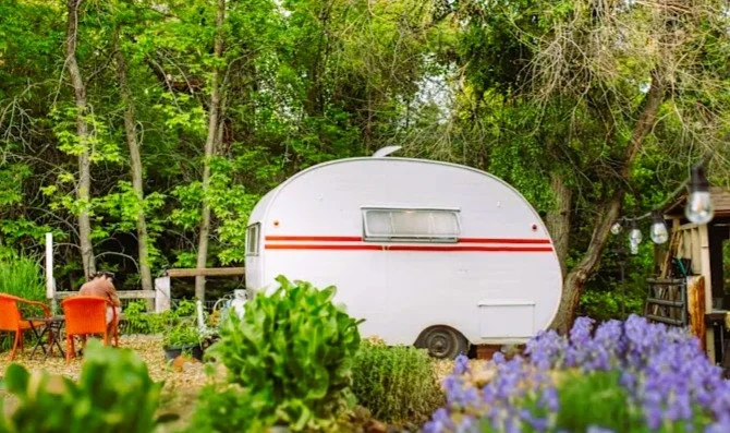 Red-Stripe Rhonda Riverside Camper surrounded by garden beds, flowers, and trees