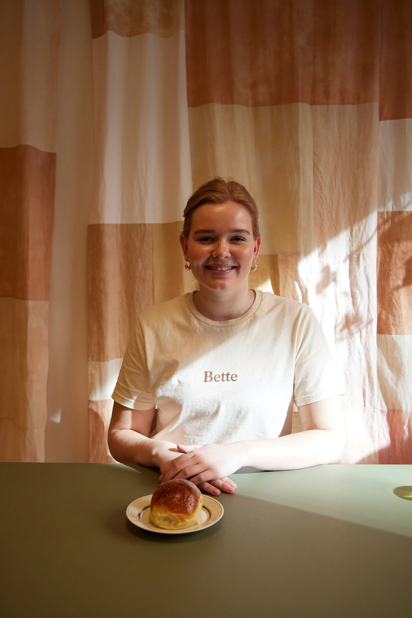 Person i en hvid T-shirt med teksten "Bette" sidder ved et bord med en bolle på en tallerken foran. Baggrunden er lys med stribet gardin.