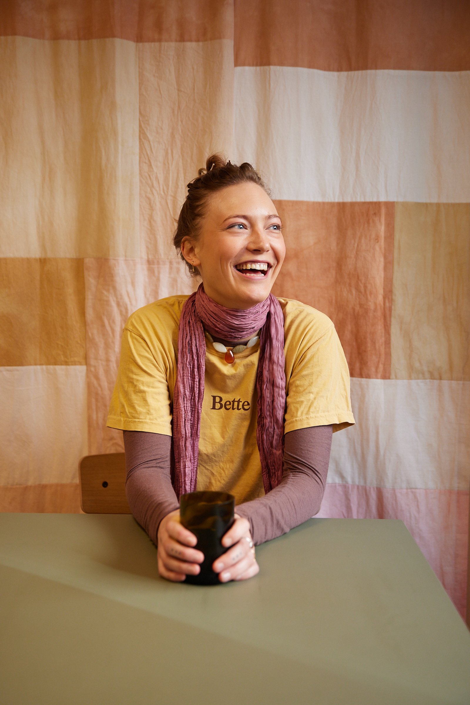 Kvinde griner og sidder ved et bord, iført gul t-shirt og lyserødt tørklæde, med en kop i hænderne.