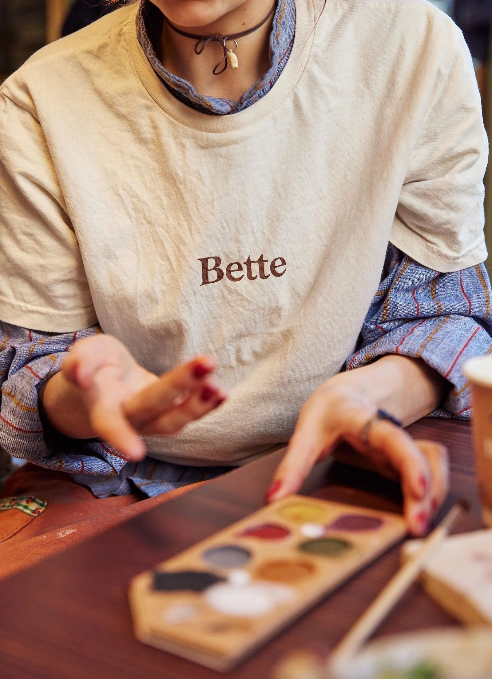 En person iført en cremefarvet t-shirt med ordet "Bette" påtrykt, sidder ved et bord og holder en farvepalette. Bordet har forskellige ting på sig, herunder en kop.