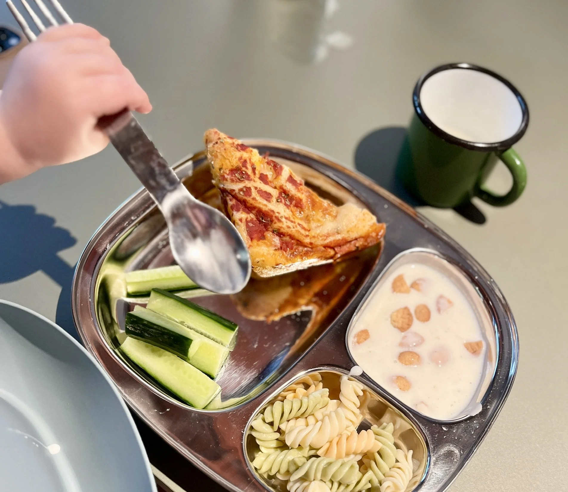 Barnearm holder en ske ved en tallerken med pizza, agurkestrimler, pasta og en skål med yoghurt eller dessert. En grøn kop står ved siden af.