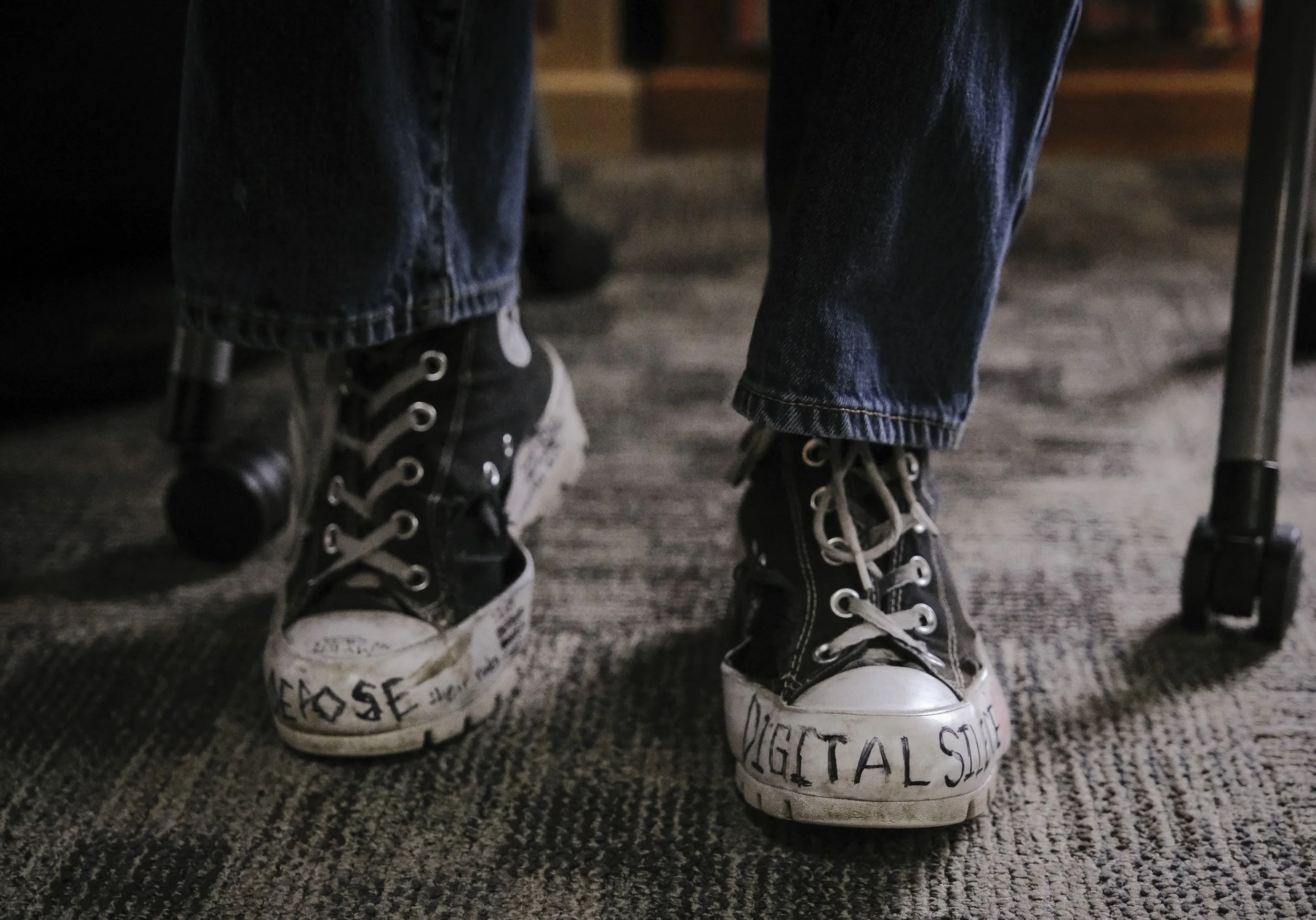 Jose Usher wears a pair of black converse shoes scribbled with political messages like, "Digital Silence", "Deny, Defend, Depose", "Eat the rich, feed the poor", and "Kamala Harris 2028."
