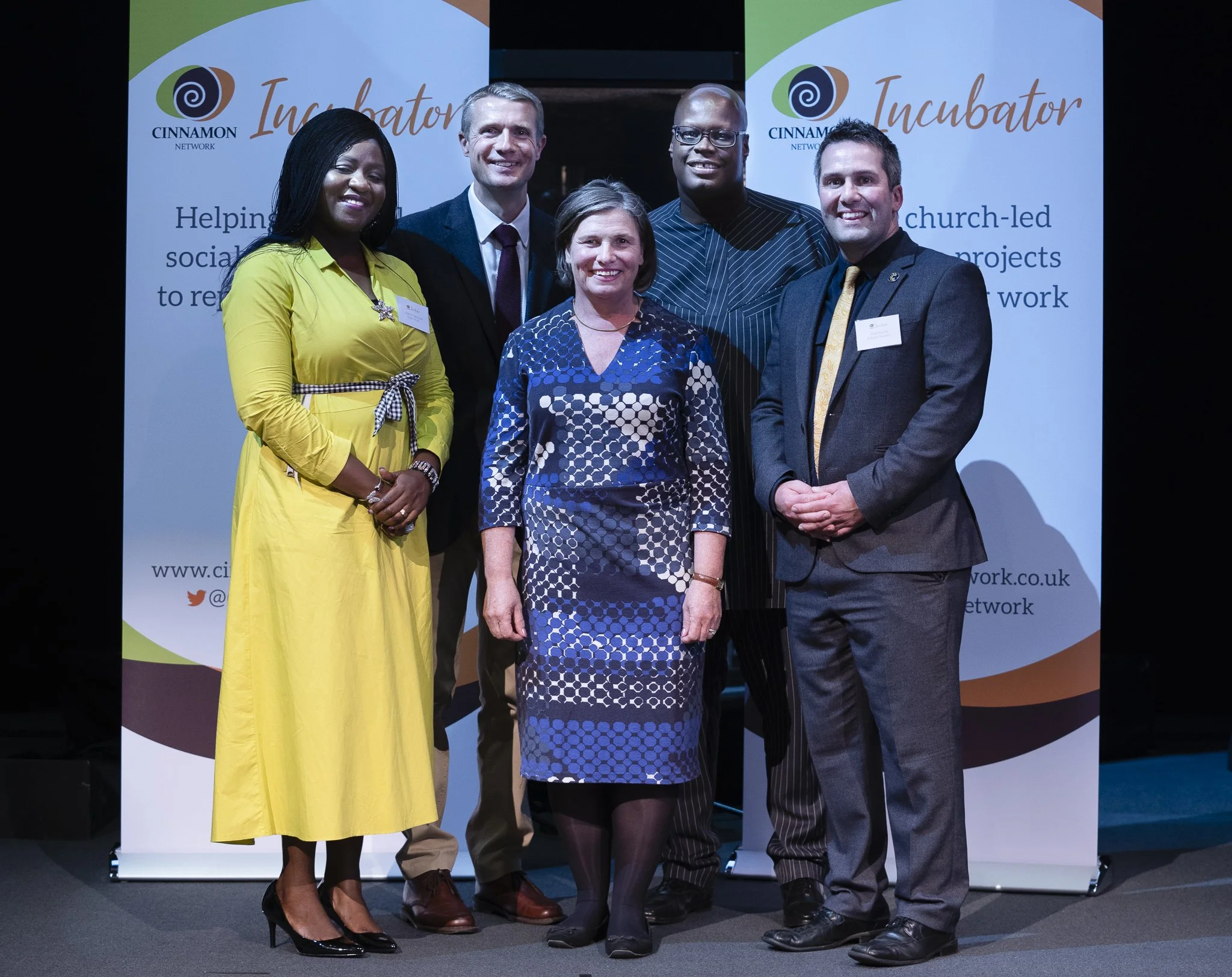 Group of six professionally dressed adults standing on stage in front of banners for the Cinnamon Network Incubator event.