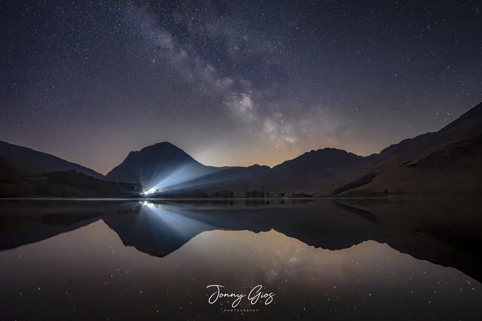 Nighttime landscape of a mountain reflected in a lake with starry sky and the Milky Way visible, with mountains in the background and a bright light source near the horizon.