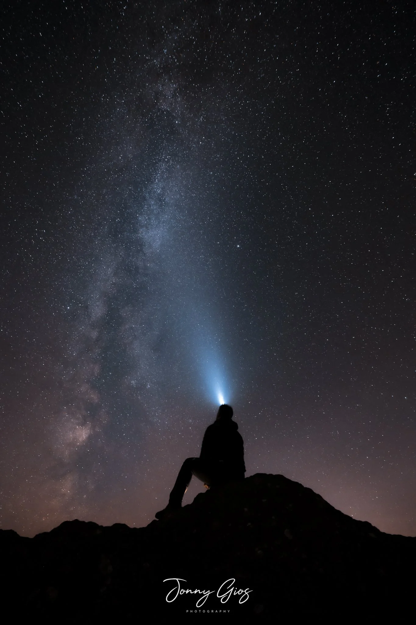 Silhouette of a person sitting on a rock at night, looking up at the star-filled sky and the Milky Way galaxy, with a light shining from their forehead.