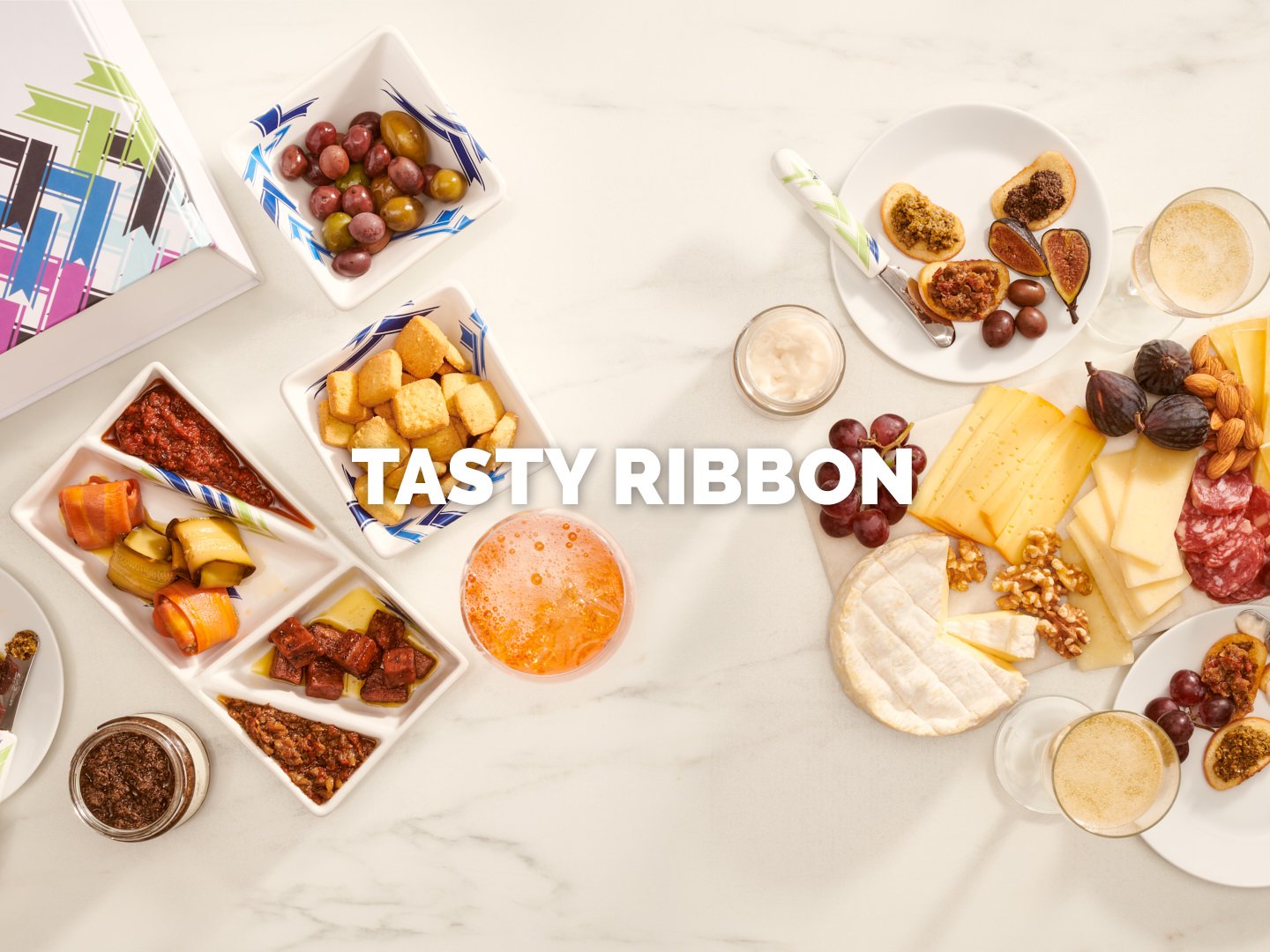 A beautifully arranged gourmet food photography spread for Tasty Ribbon. The composition showcases a selection of artisanal cheeses, charcuterie, marinated vegetables, olives, and gourmet spreads, elegantly presented with crisp crackers and a glass o