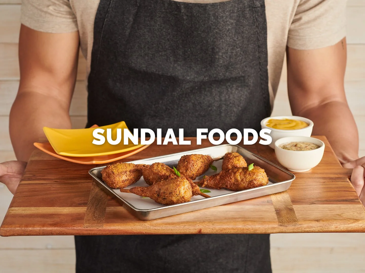 A compelling food photography shot showcasing a tray of crispy, golden-brown plant-based drumsticks from Sundial Foods. The warm, rustic presentation features a chef holding the dish on a wooden board, accompanied by vibrant dipping sauces. The compo