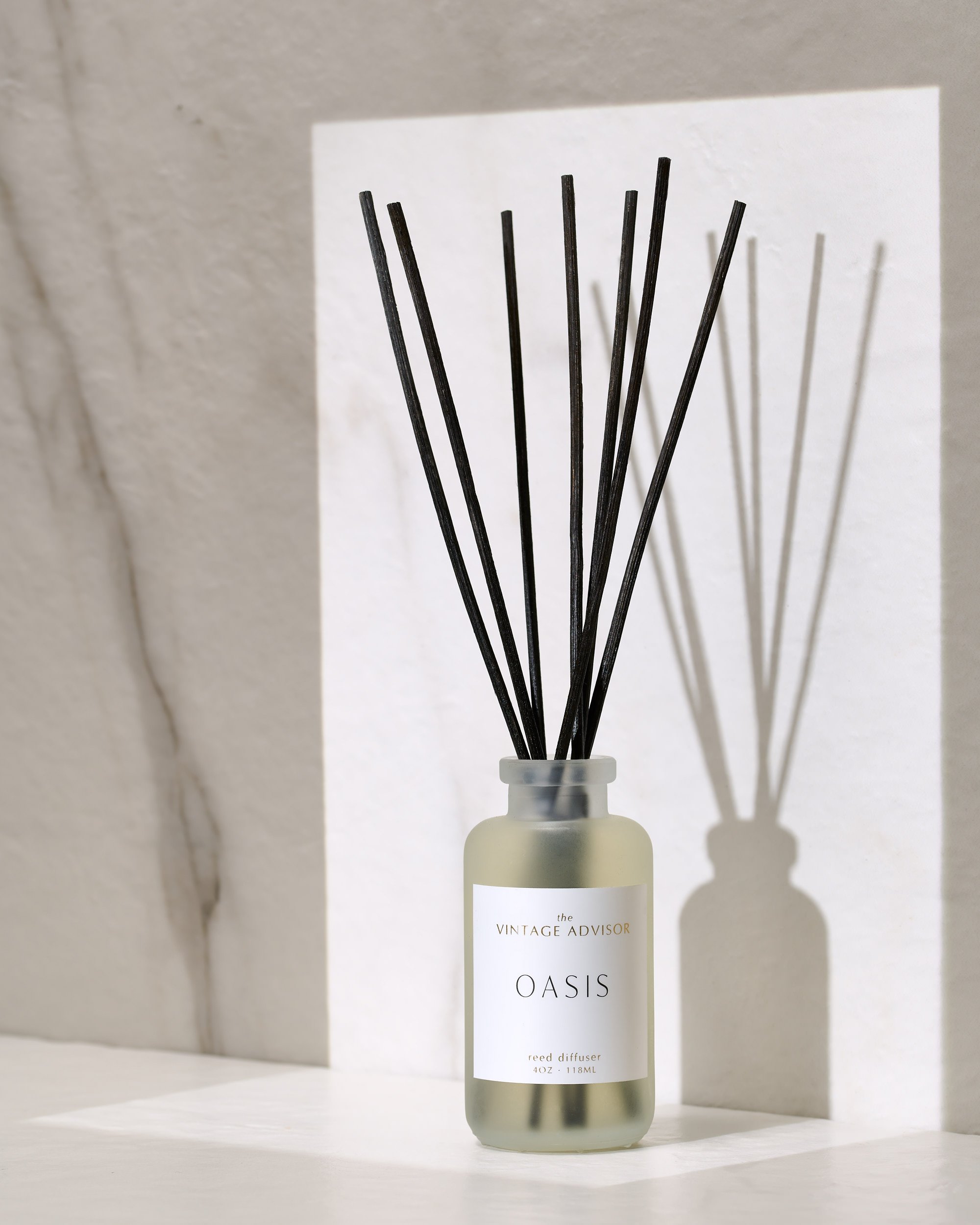 A frosted glass reed diffuser bottle labeled 'OASIS' by Vintage Advisor with black reed sticks inside, casting their shadow on a white wall.
