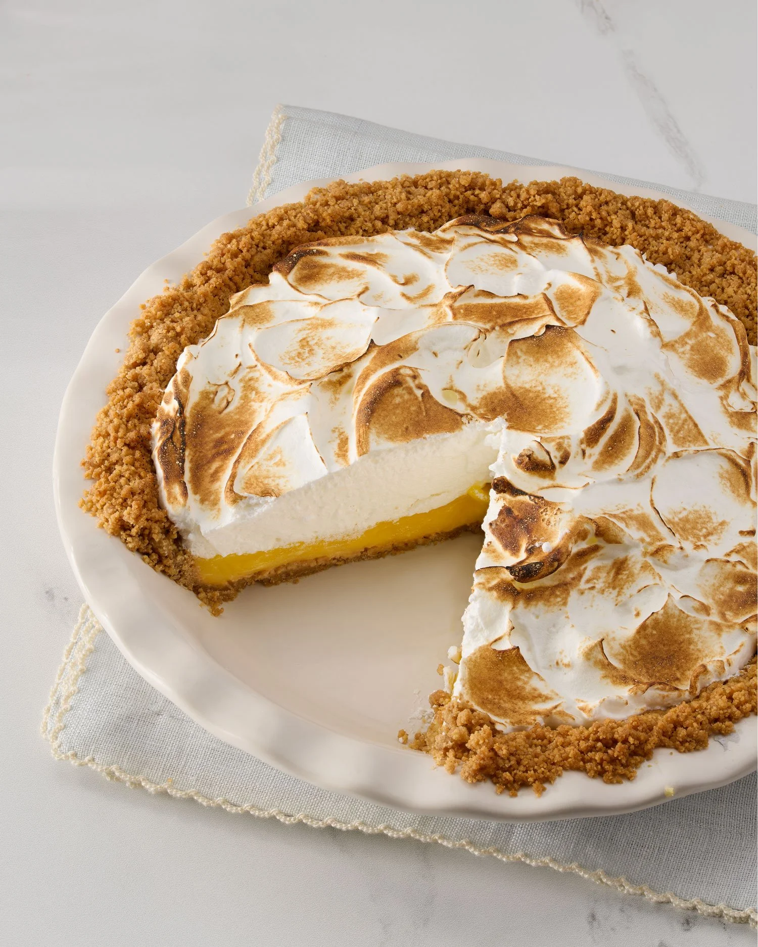 A lemon meringue pie with toasted meringue topping on a white plate, with a slice removed showing lemon filling and crust.