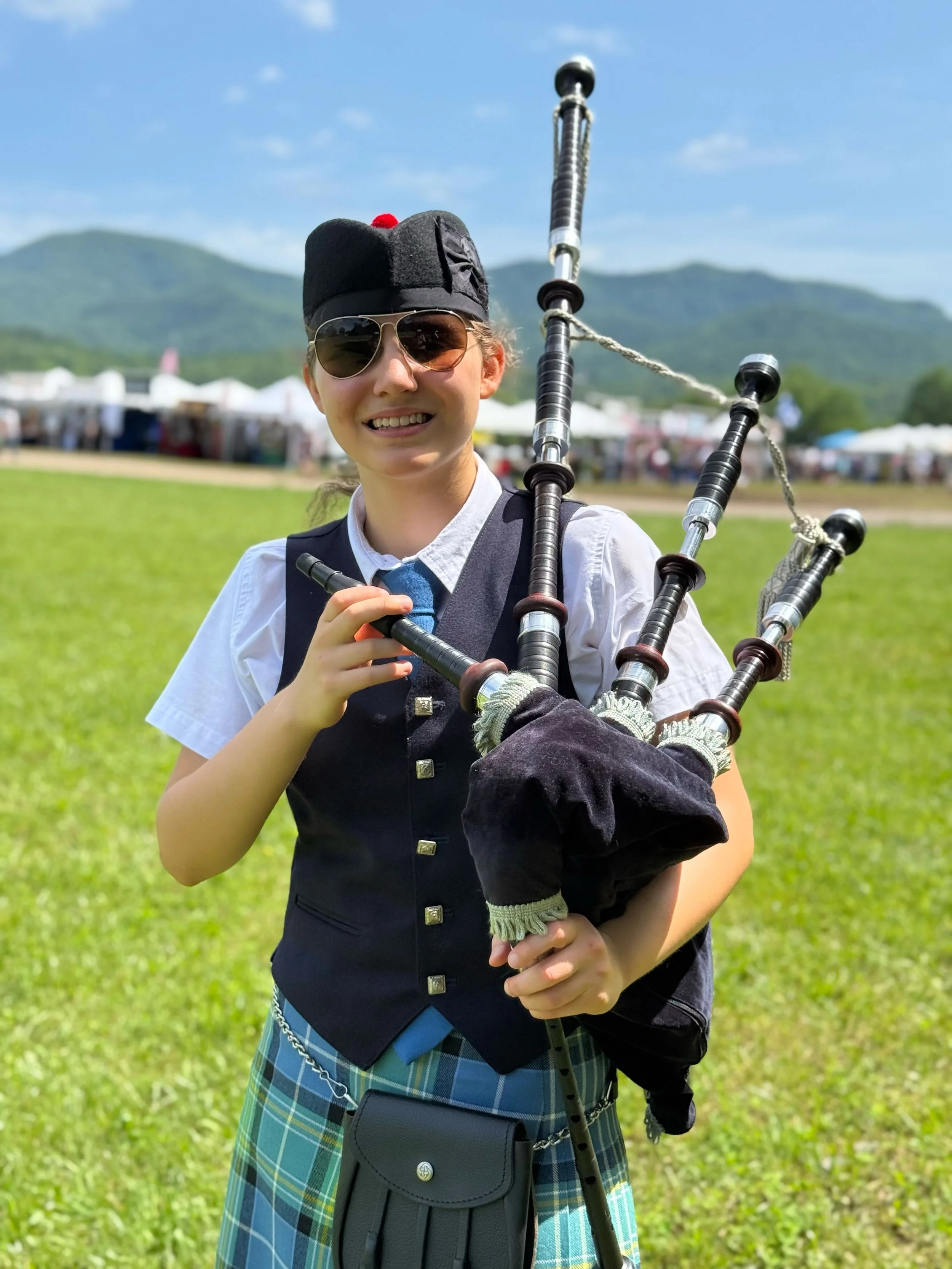 Two young men in uniform, with one carrying a bag and a sword, outdoors during daytime, with trees and a white vehicle in the background.