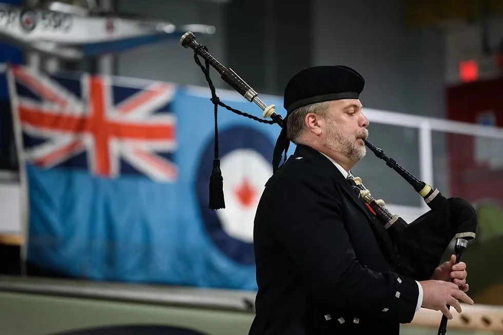 A man playing bagpipes in front of a flag that combines the British Union Jack and the Canadian maple leaf, likely indoors at a cultural event or gathering.