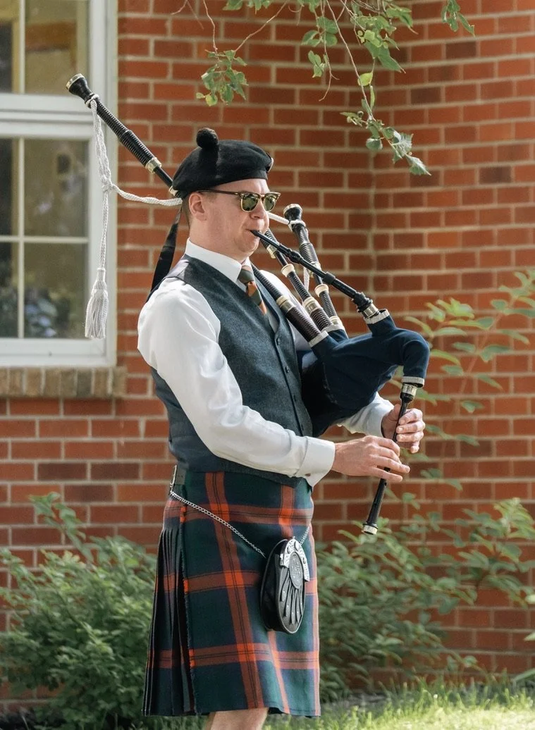 A young man wearing a black hat and blue shirt holding a large silver trophy, smiling outdoors with trees and a blurred background.