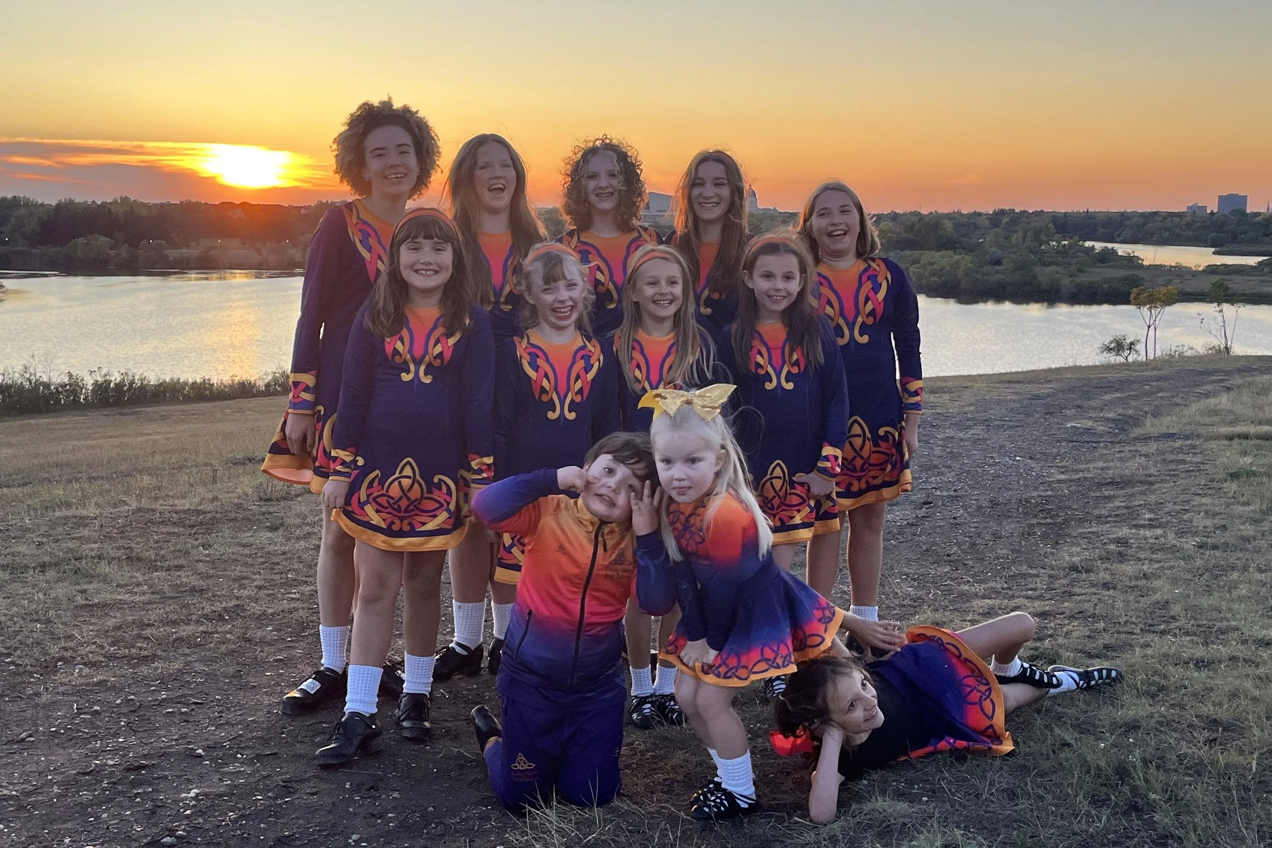 A group of young girls in colorful matching cheerleader uniforms posing outdoors at sunset near a river and trees, some standing and some sitting or lying on the ground, smiling and enjoying the moment.