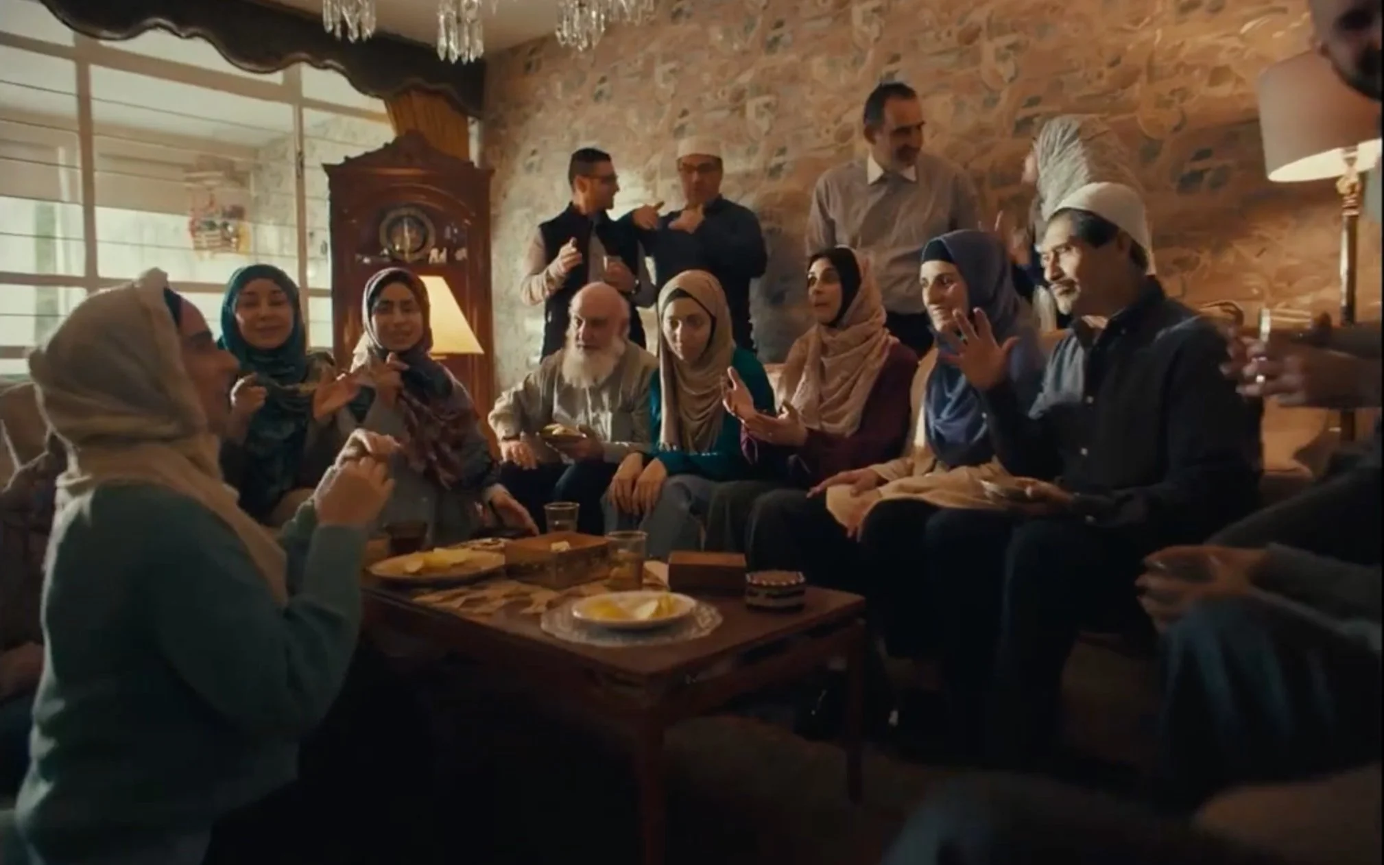 Un grupo de personas en una reunión en una sala con paredes de ladrillo, algunos con ropa tradicional, algunos en conversación y otros haciendo gestos. Hay comida en la mesa central.