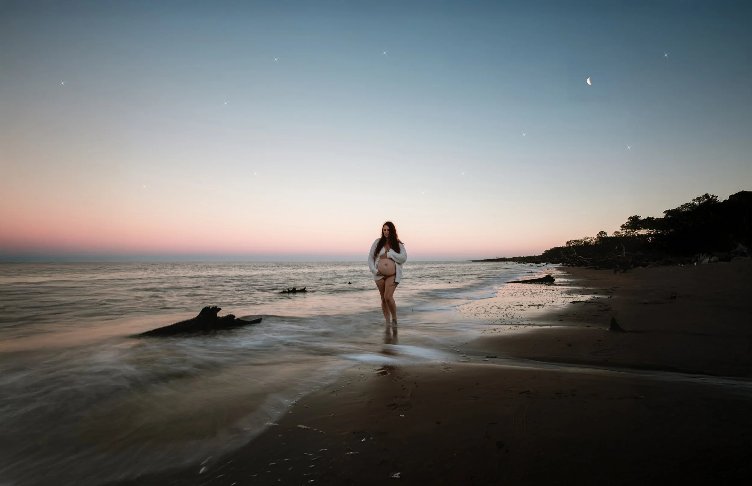 A pregnant woman standing on a beach during sunset, with the sky showing stars and a crescent moon, gentle waves at her feet, and a silhouette of trees in the background.