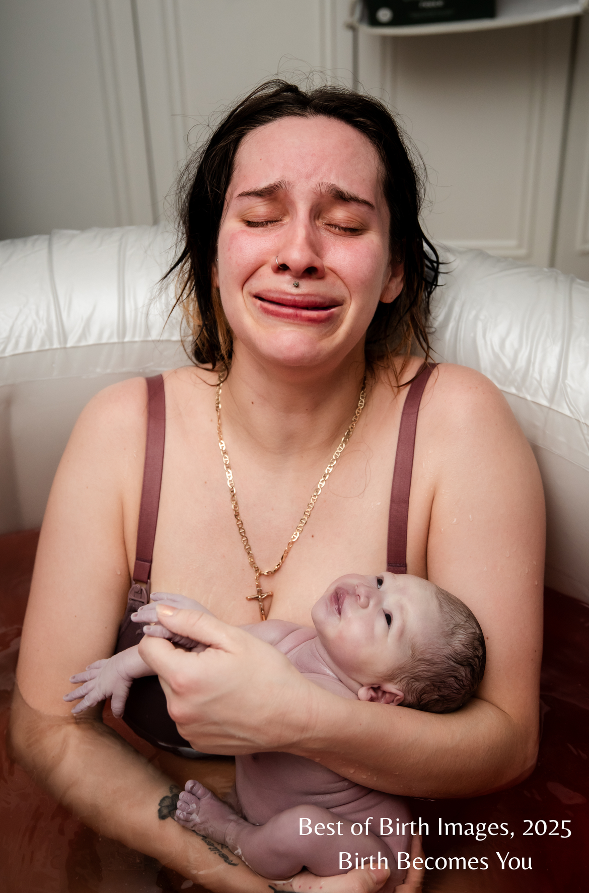 A woman with short dark hair, crying tears of joy while holding a newborn baby during a water birth in a birthing pool, with a white background and a caption that reads 'Best of Birth Images, 2025 Birth Becomes You.'