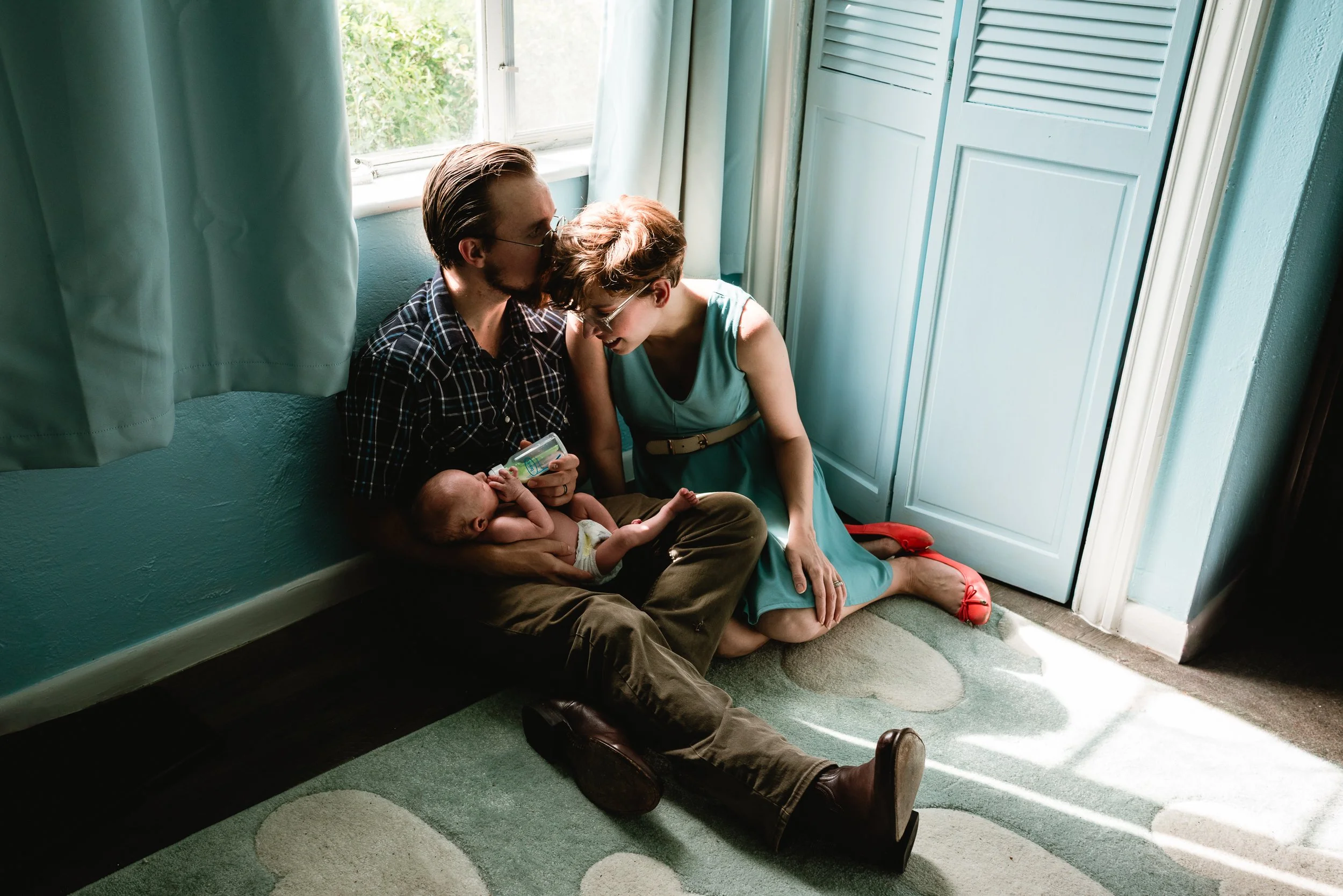 A man and woman sit on the floor next to a window, holding a baby and bottle, with sunlight shining in.
