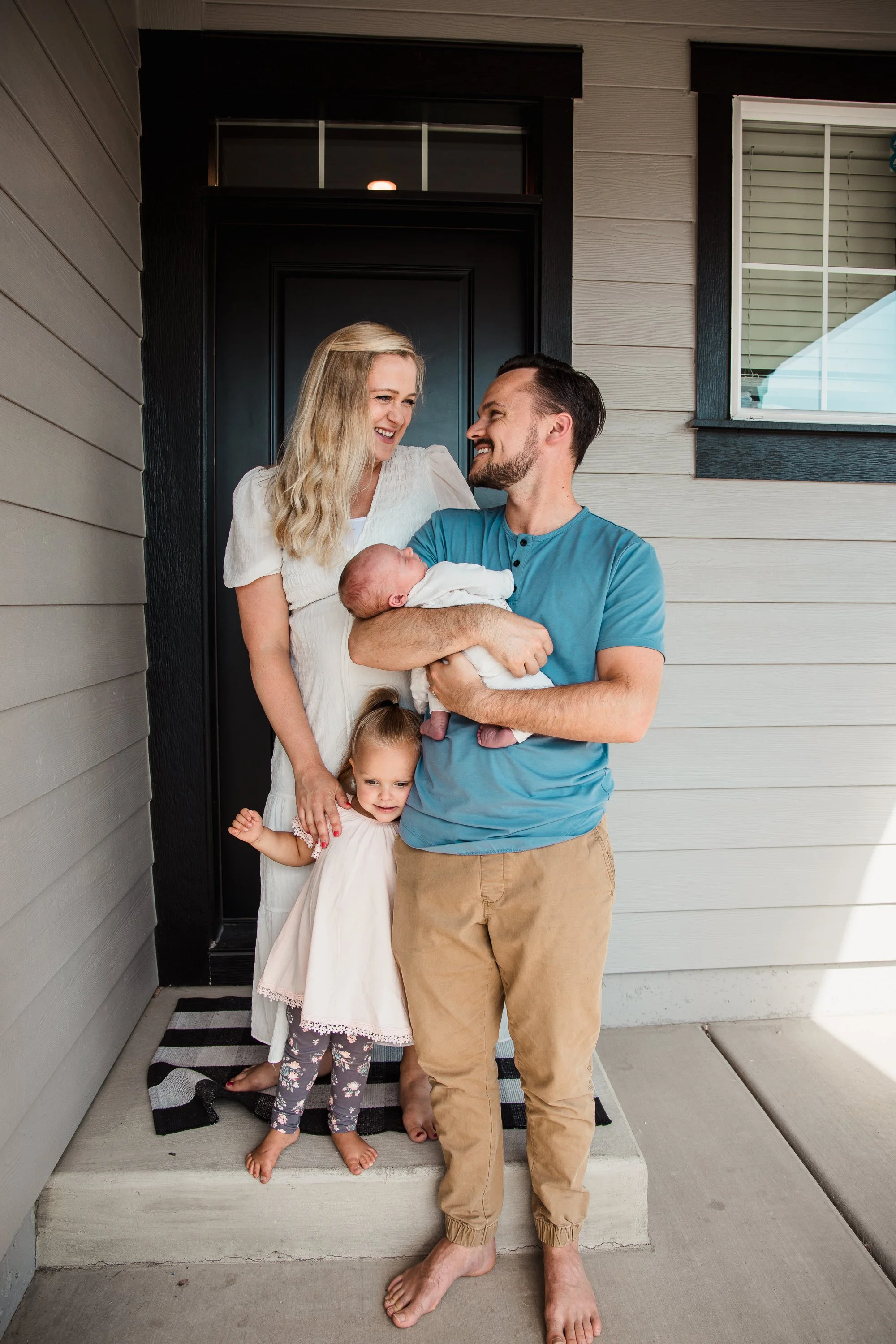A family of four standing on a porch in front of a house, with a woman, a man, a newborn baby, and a young girl. The woman with blonde hair is wearing a white dress, and the man with dark hair and a beard is wearing a blue t-shirt and tan pants. The woman is holding the sleeping newborn baby dressed in white, and the young girl is standing in front of them wearing a white dress and floral leggings. The family appears happy and smiling.