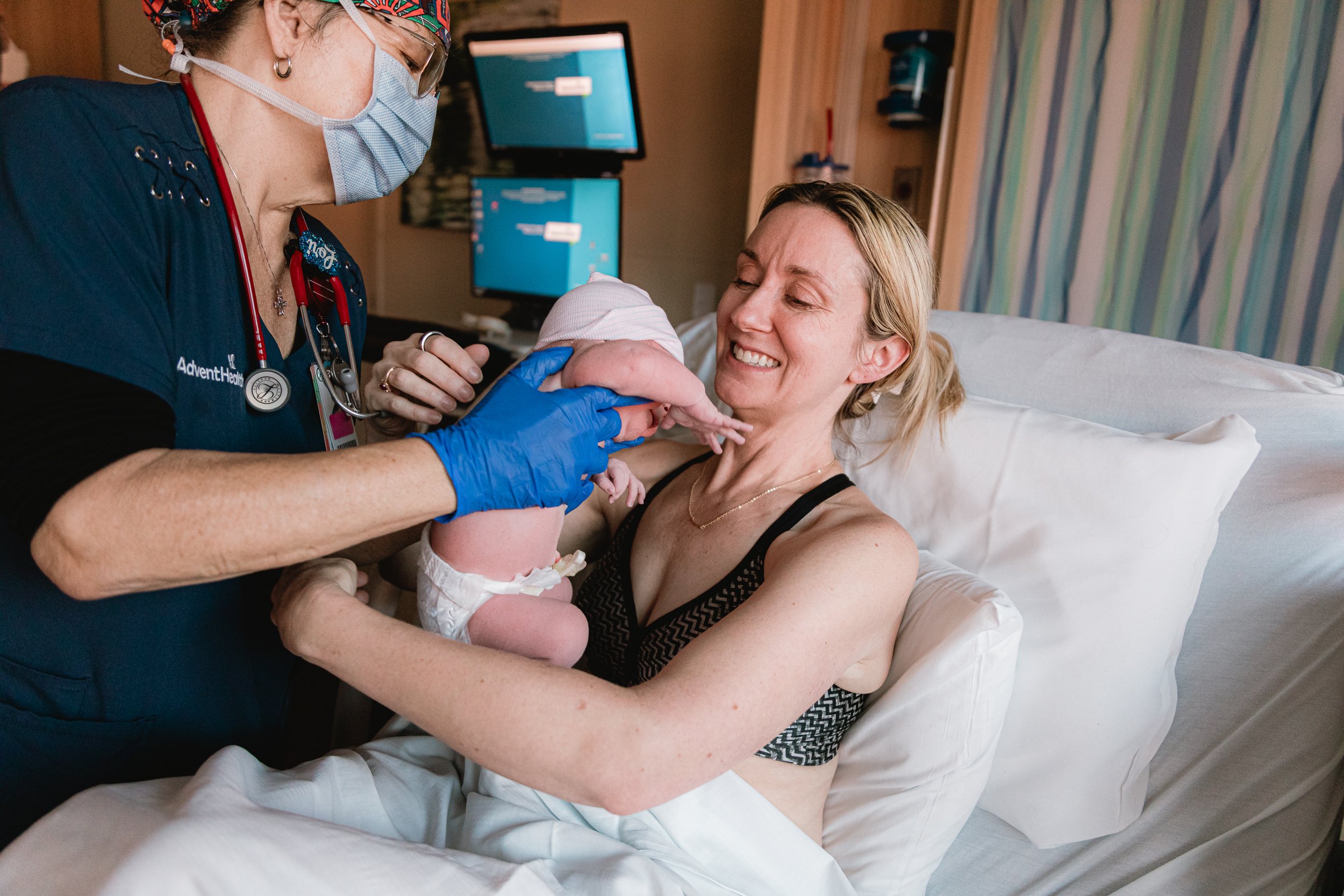 A woman in labor holding her newborn baby with a nurse assisting, in a hospital room.