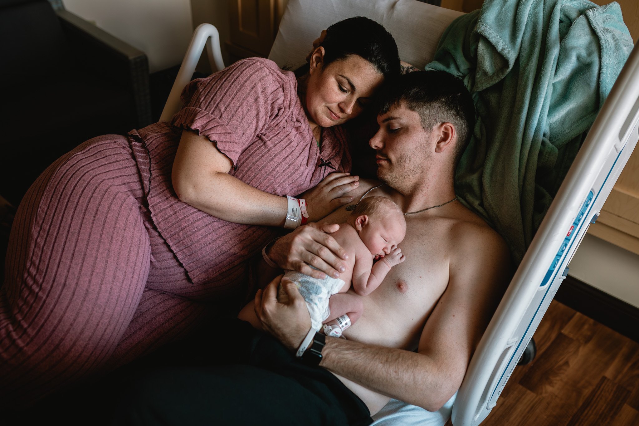 A woman in hospital pajamas gently holding a newborn baby on a hospital bed from a side angle. A man, lying shirtless with a hospital wristband and medical implant, is closely observing the baby. The scene is in a hospital room with wooden floors.