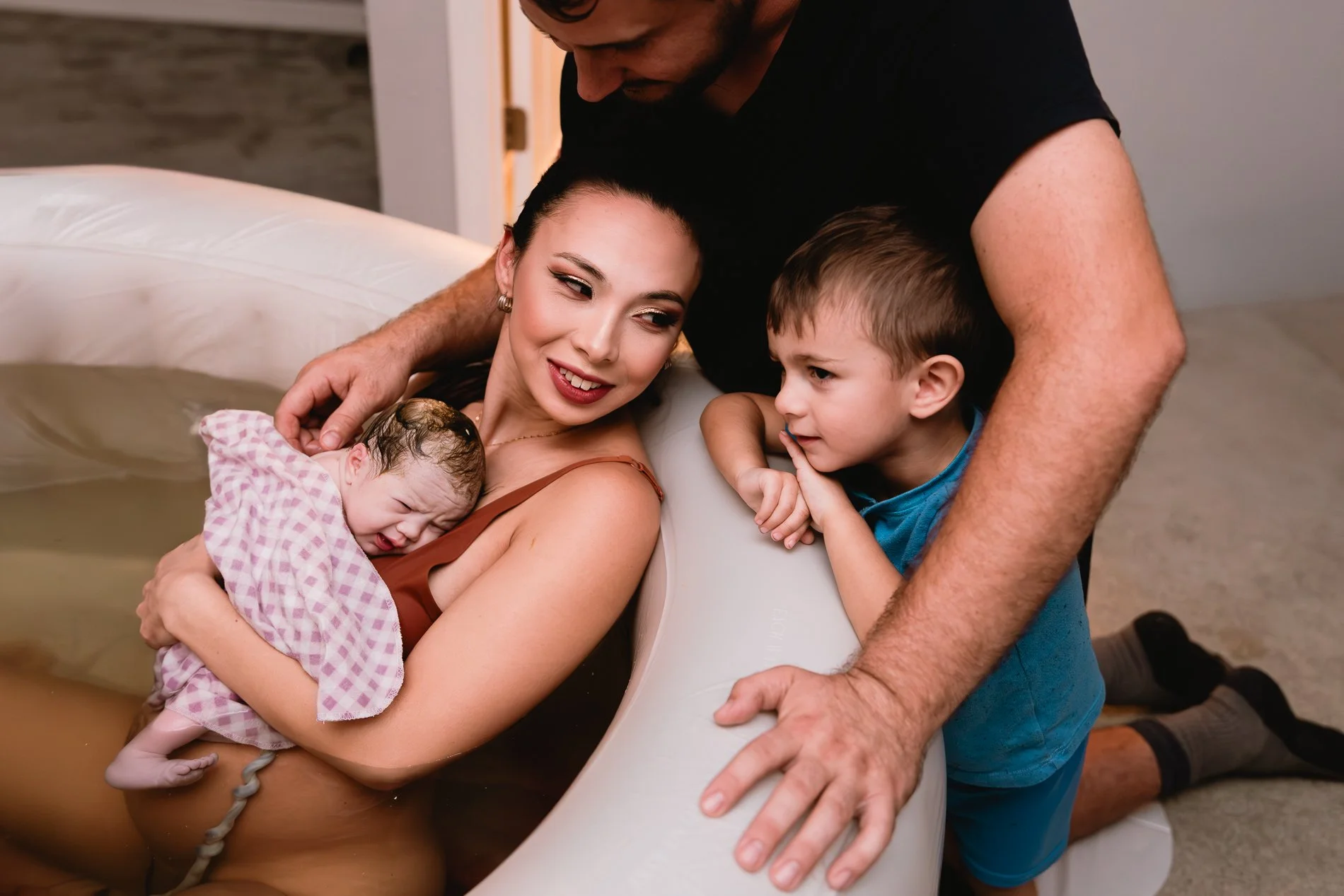 A woman giving birth in a birthing pool, cradling her newborn baby. A man and a young boy are leaning over the pool, watching the baby and supporting the woman.