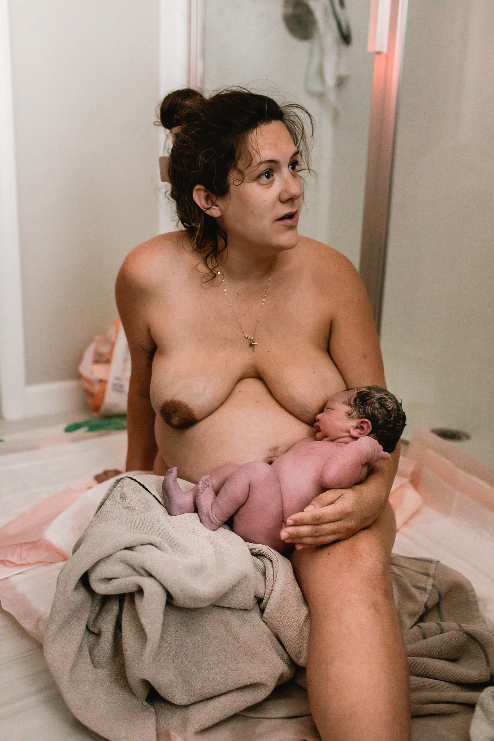 A woman with dark hair, partially dried, holding a newborn baby with dark curly hair against her chest, sitting on the floor of the bathroom with a shower door in the background.