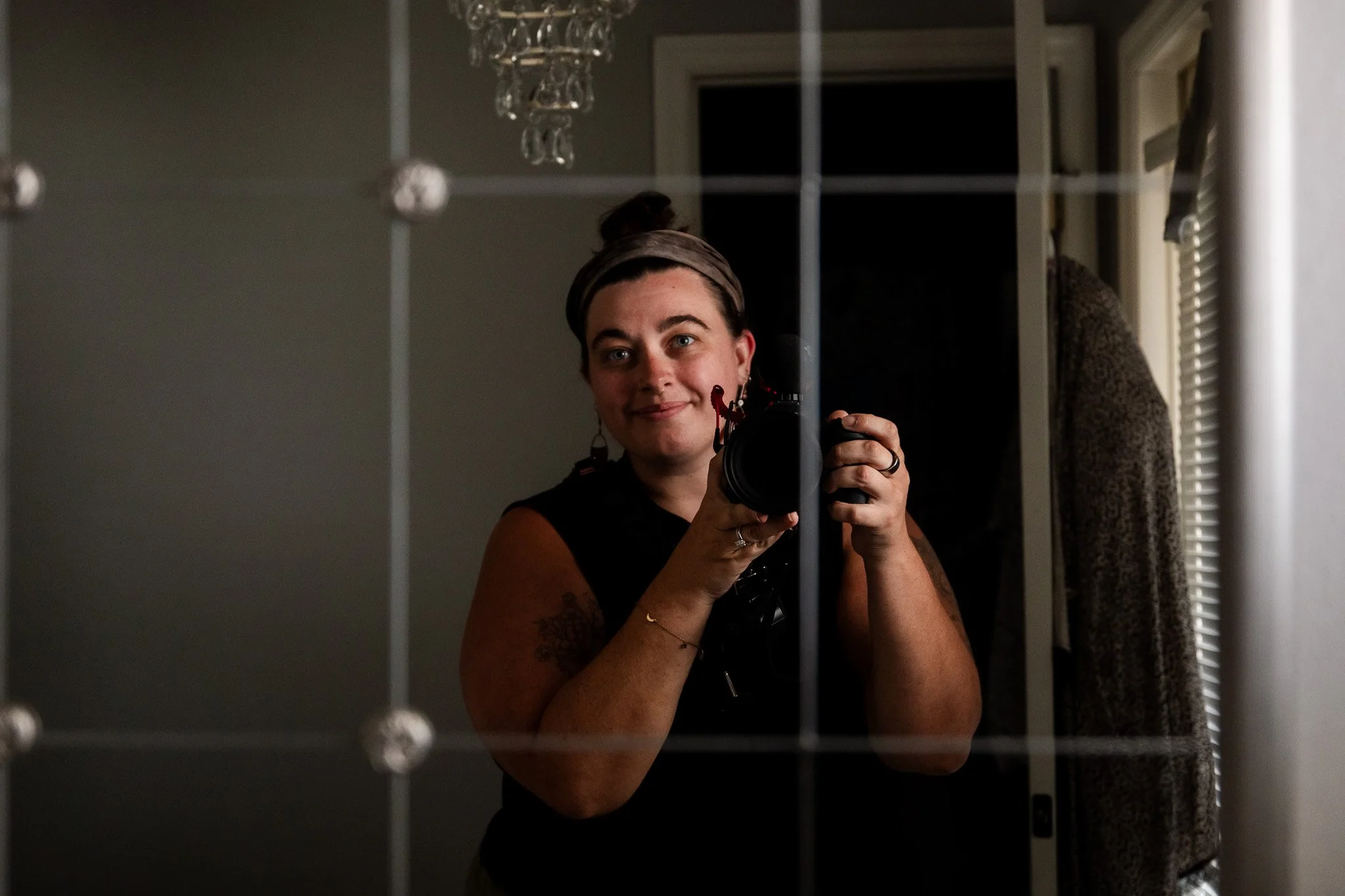 A person with dark hair tied up, holding a camera, taking a mirror selfie in a dimly lit room with a chandelier and window blinds.