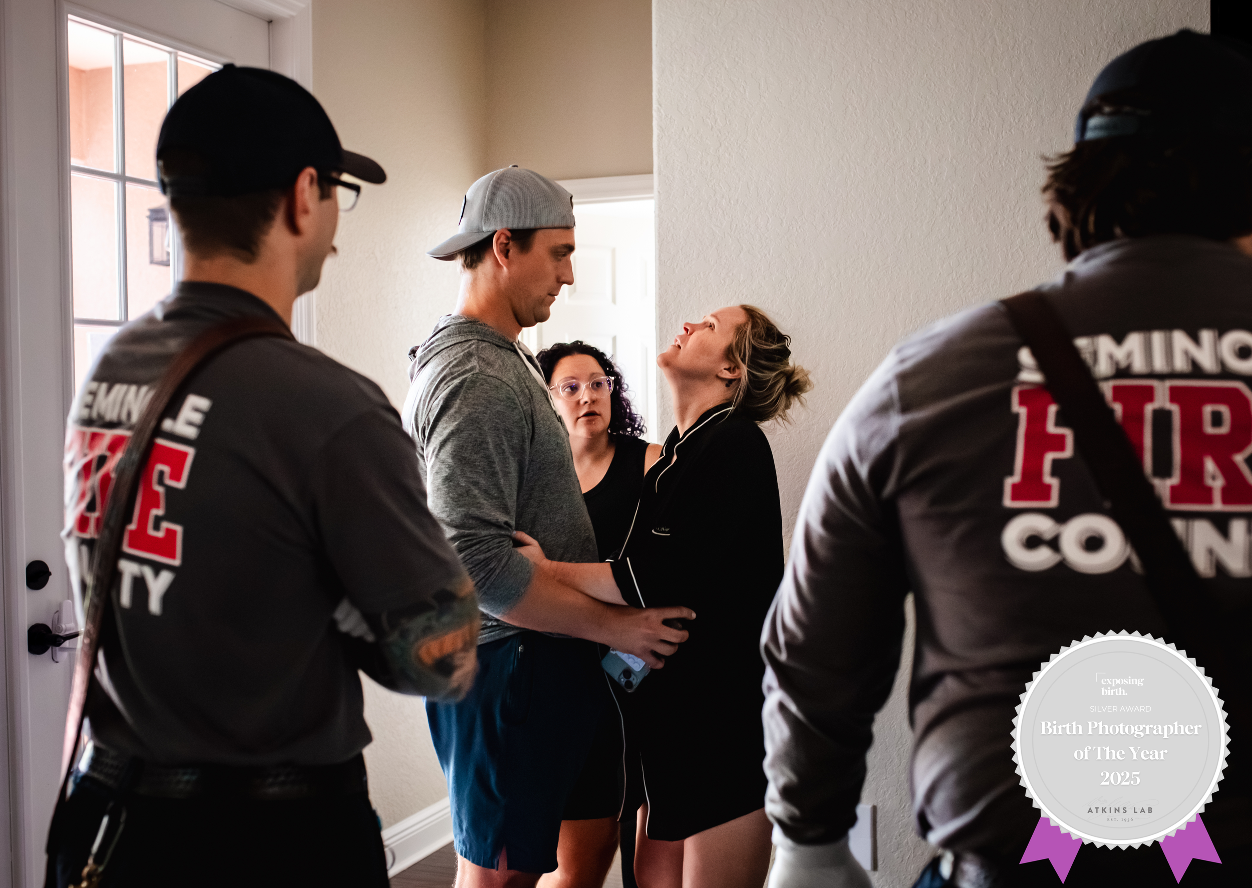 A woman appears to be in active labor at home, with her partner and emergency responders present, as she leans back and screams while another responder looks surprised. The scene shows a doorway and other emergency personnel in shirts labeled 'FIRE' 