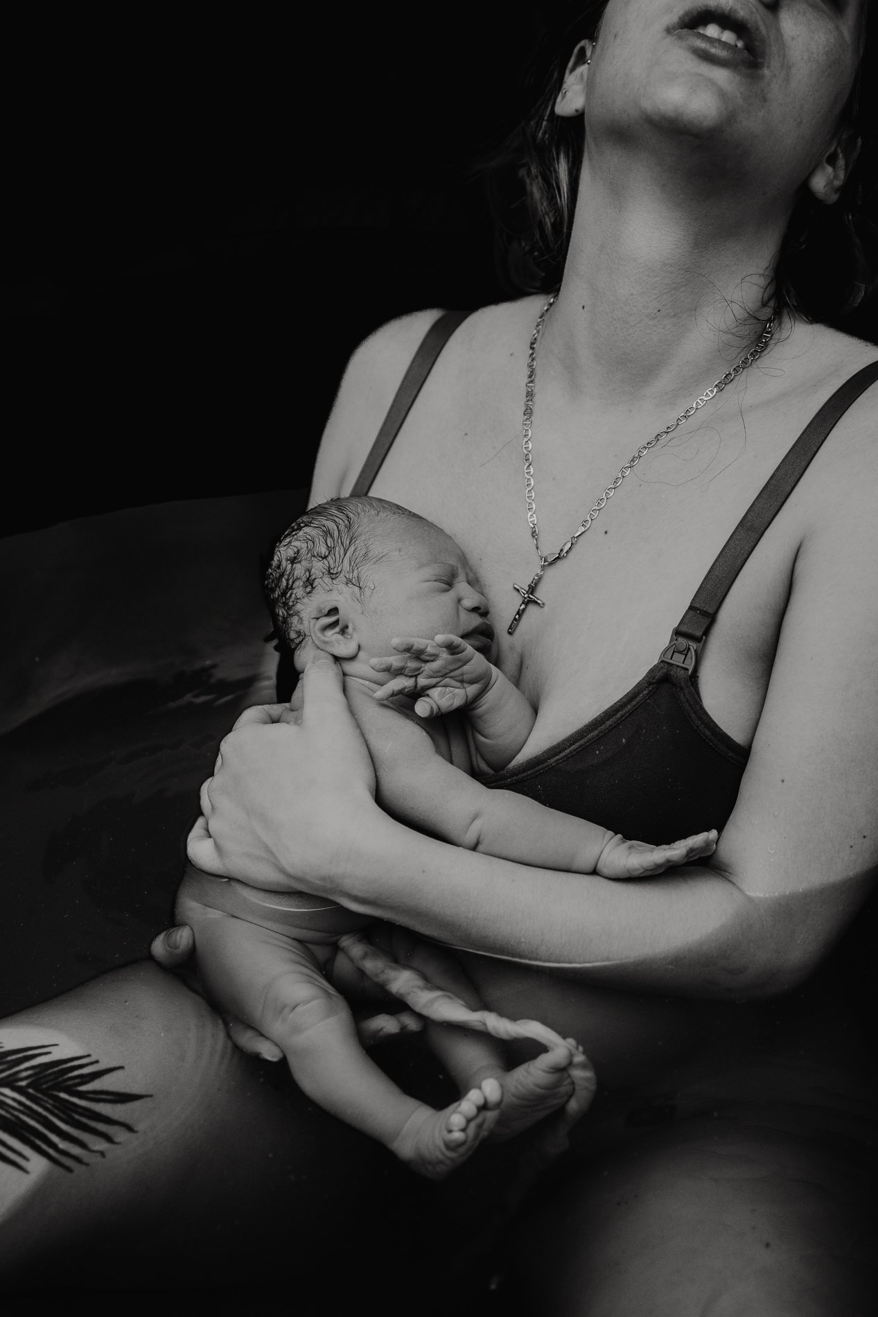 A woman is holding a newborn baby during a water birth. The woman wears a necklace with a cross.