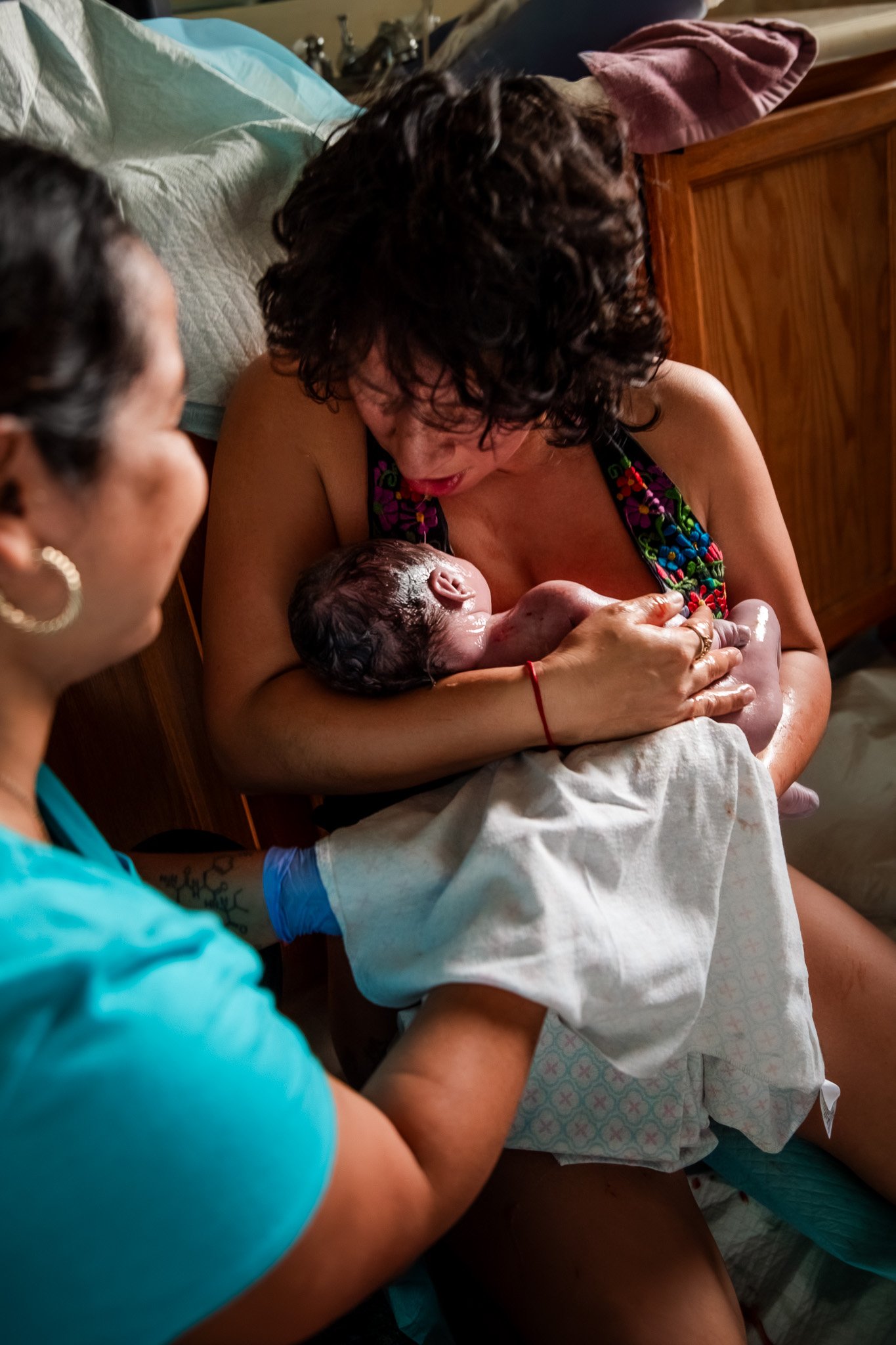 A woman, possibly the mother, holds her newborn baby, cradling the infant close to her chest as they bond. A nurse or medical professional, wearing blue scrubs, assists in the birthing process in a hospital room, with another woman observing nearby.
