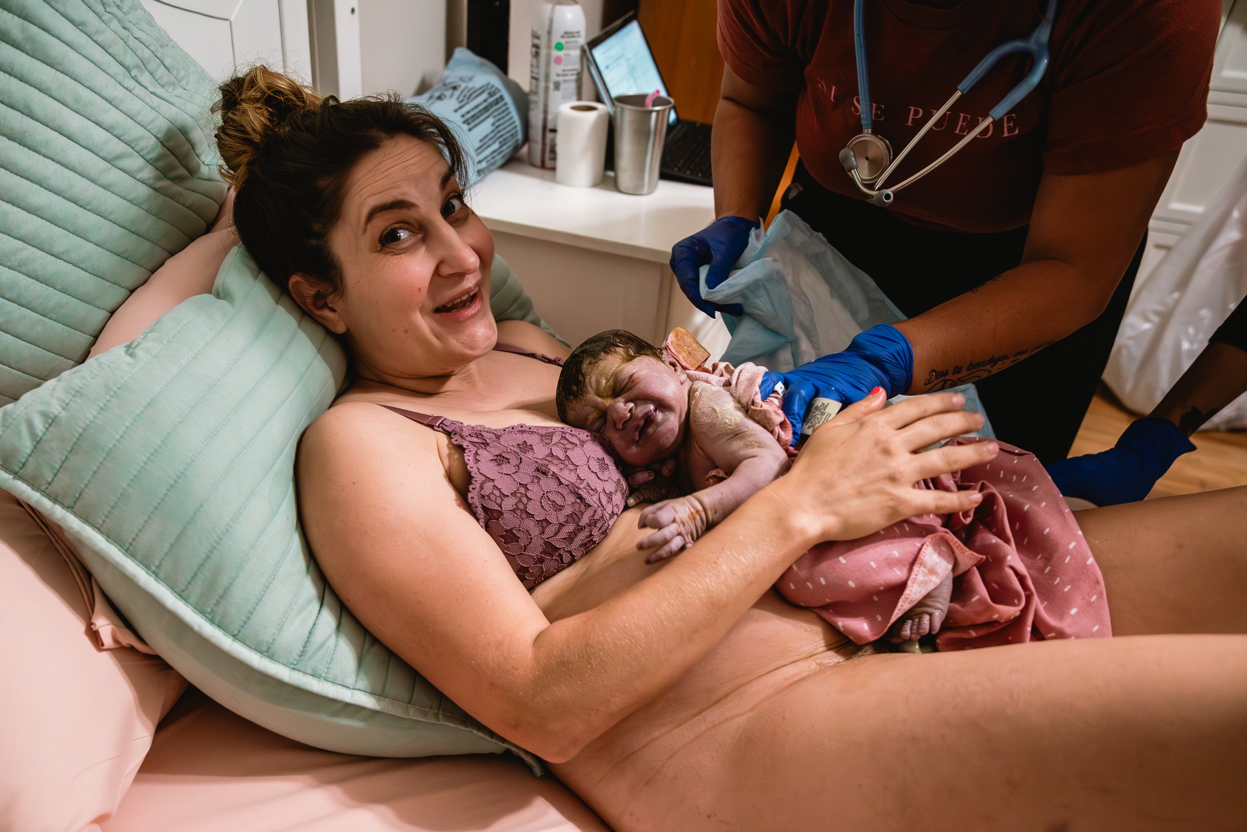 New mother holding her newborn baby right after childbirth at home, with midwife assisting.