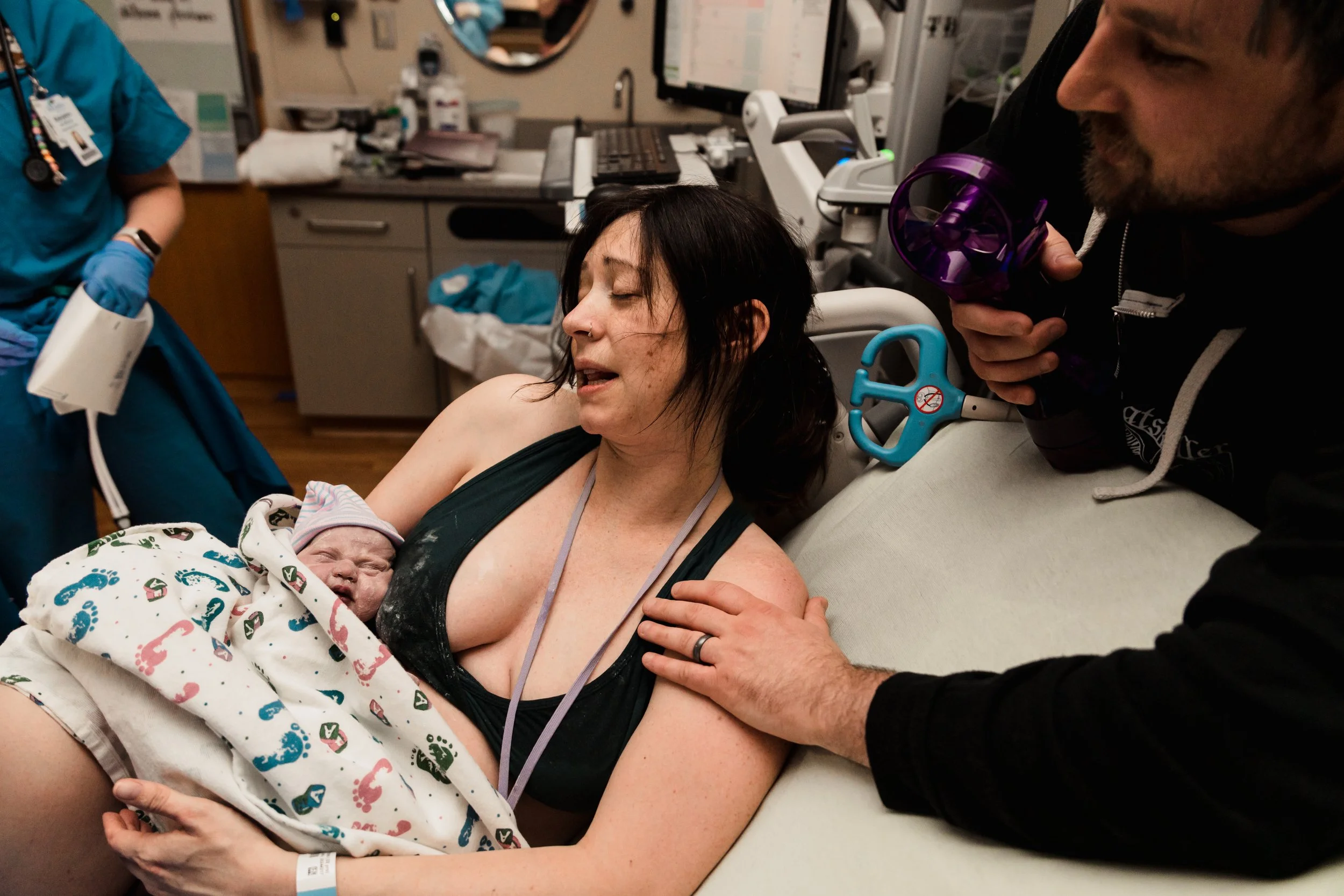 A woman holding a newborn baby in a hospital room, with a support person nearby, while a healthcare worker adjusts medical equipment.